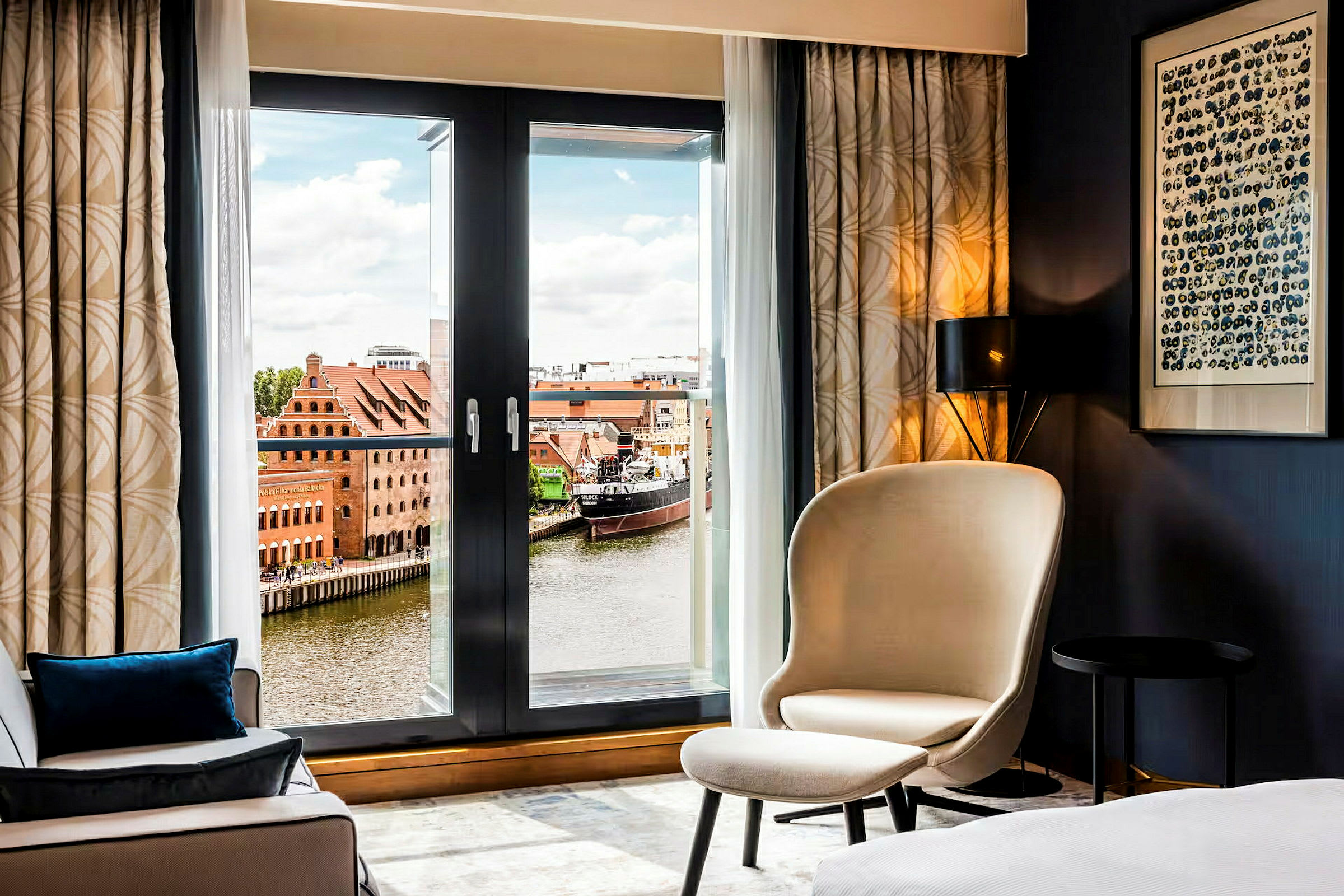 Ett hotellrum med en fåtölj och utsikt över en flod med byggnader och en båt utanför. (AI-genererad text)