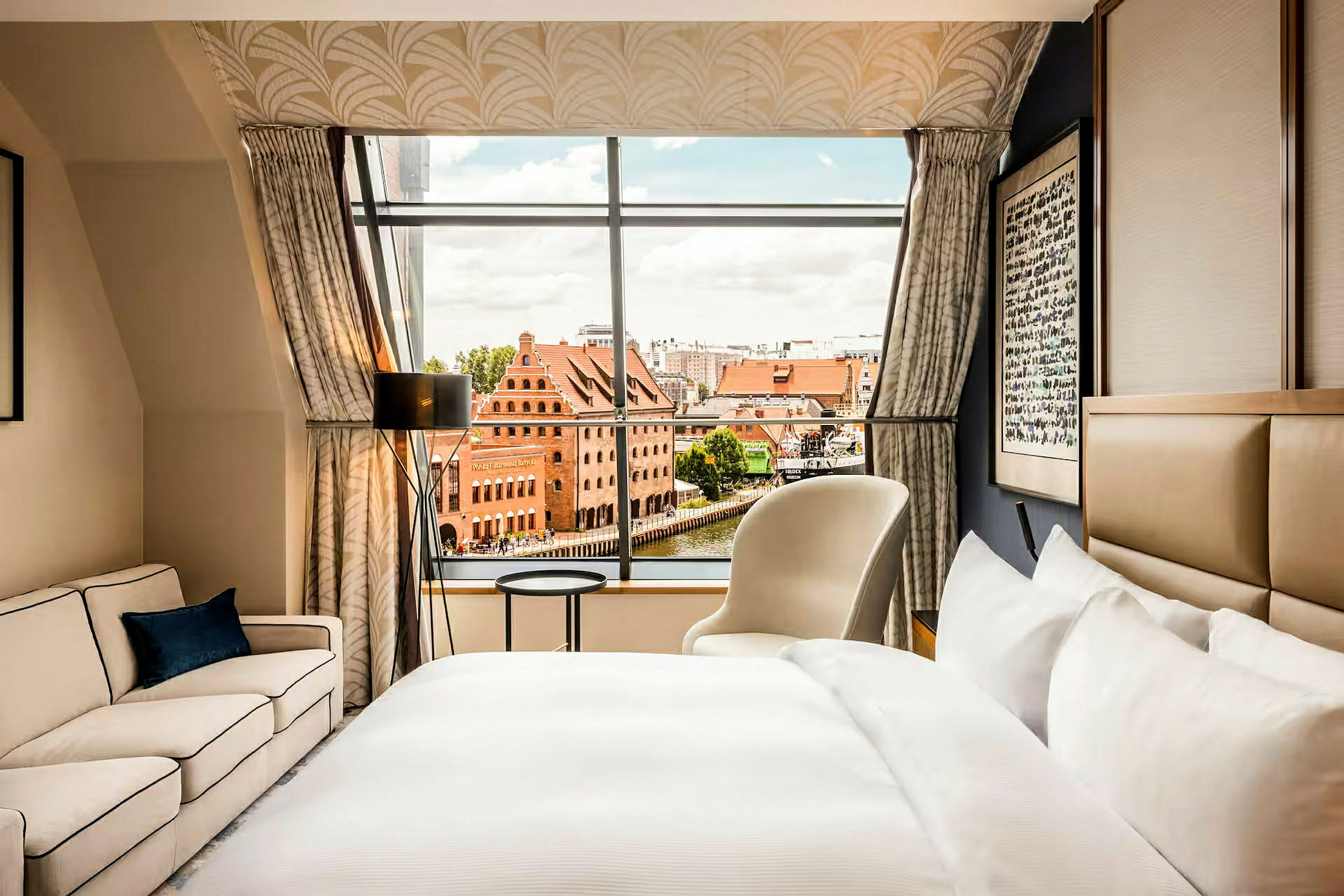 Ett hotellrum med en säng, soffa och fåtölj, samt ett stort fönster med utsikt över en stadsmiljö med röda tegelbyggnader. (AI-genererad text)