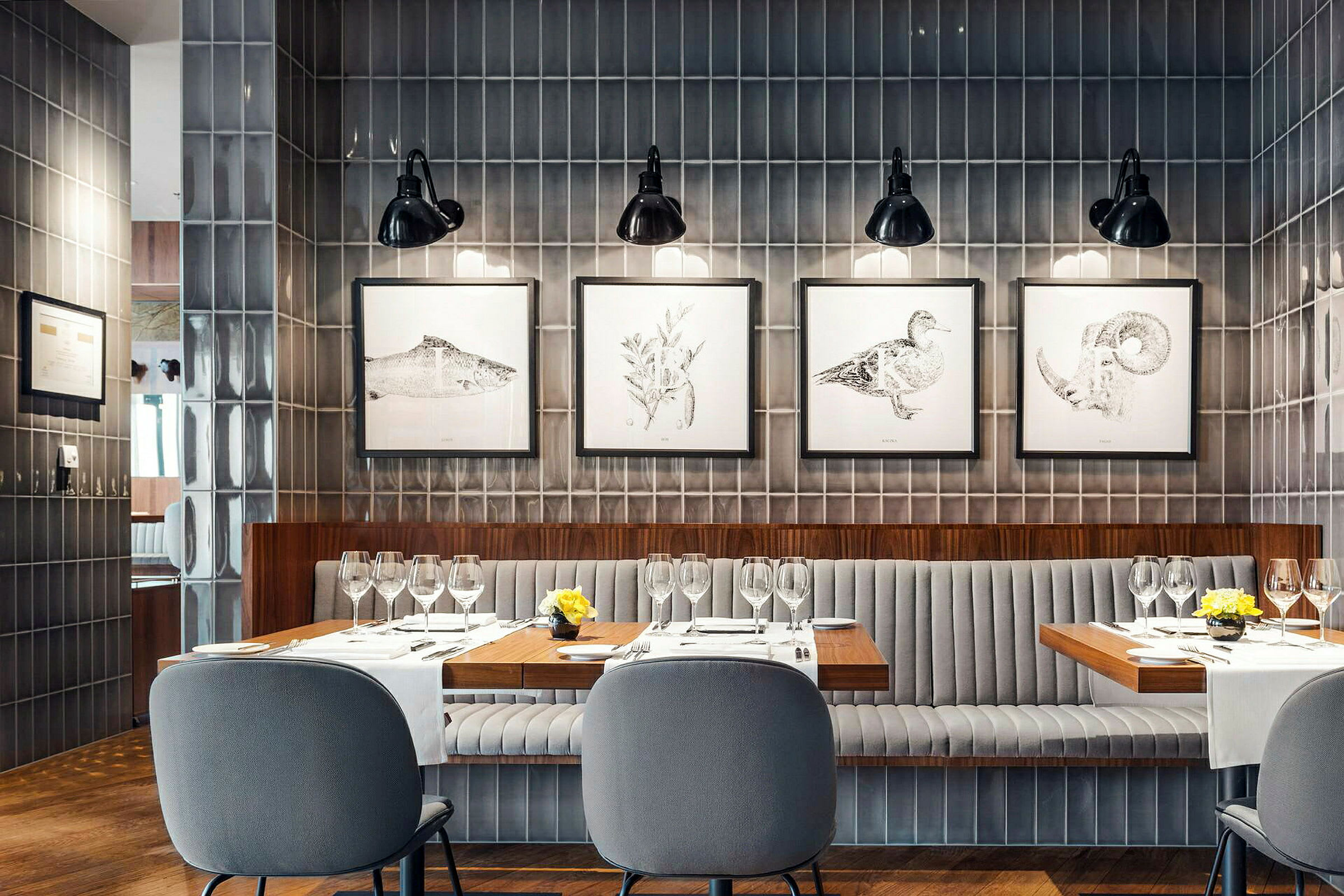 En modern restaurang med grå väggpaneler, ett bord dukat med glas och servetter samt stolar med stoppade ryggstöd. Tavlor hänger på väggen bakom. (AI-genererad text)