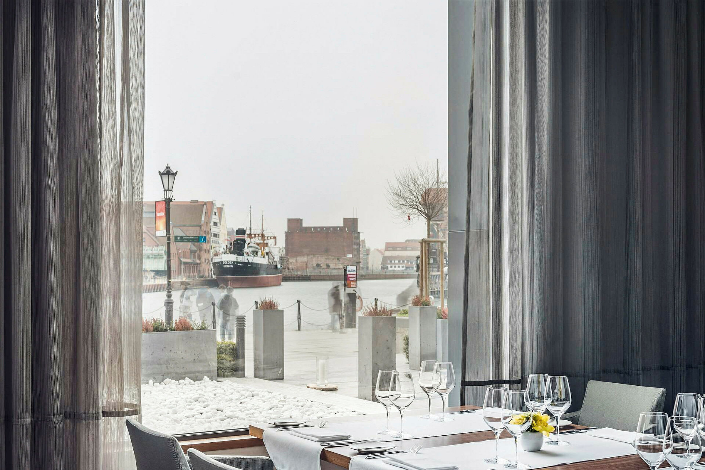 Restaurangbord bredvid ett stort fönster med utsikt över en hamn med ett fartyg. (AI-genererad text)