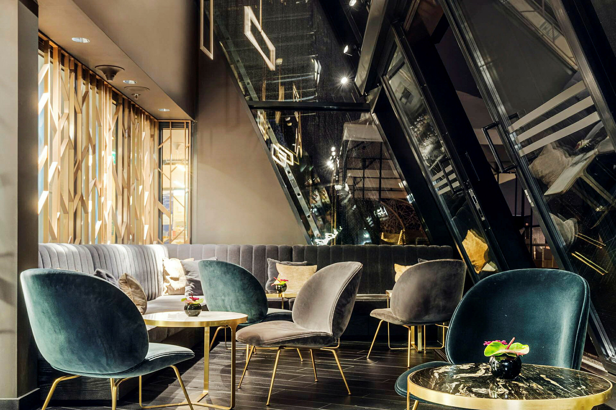 Ett modernt vardagsrum eller lounge med eleganta sammetsfåtöljer, små bord med metallram och dekorativa växter, samt stora fönster som lutar inåt. (AI-genererad text)
