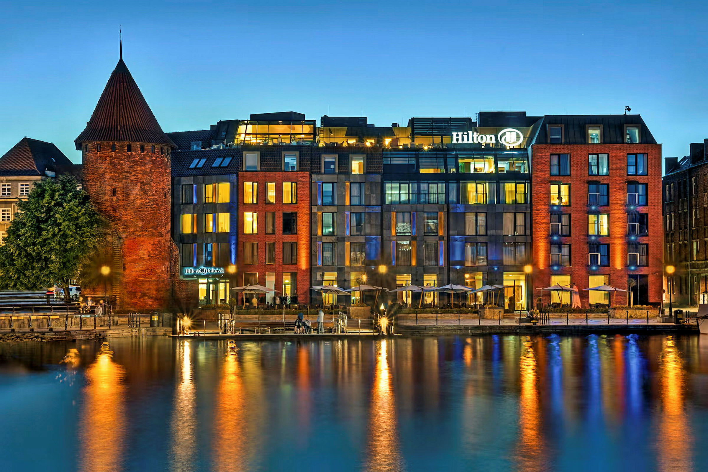 En historisk byggnad i tegel bredvid ett modernt hotell vid vattnet på kvällen. (AI-genererad text)