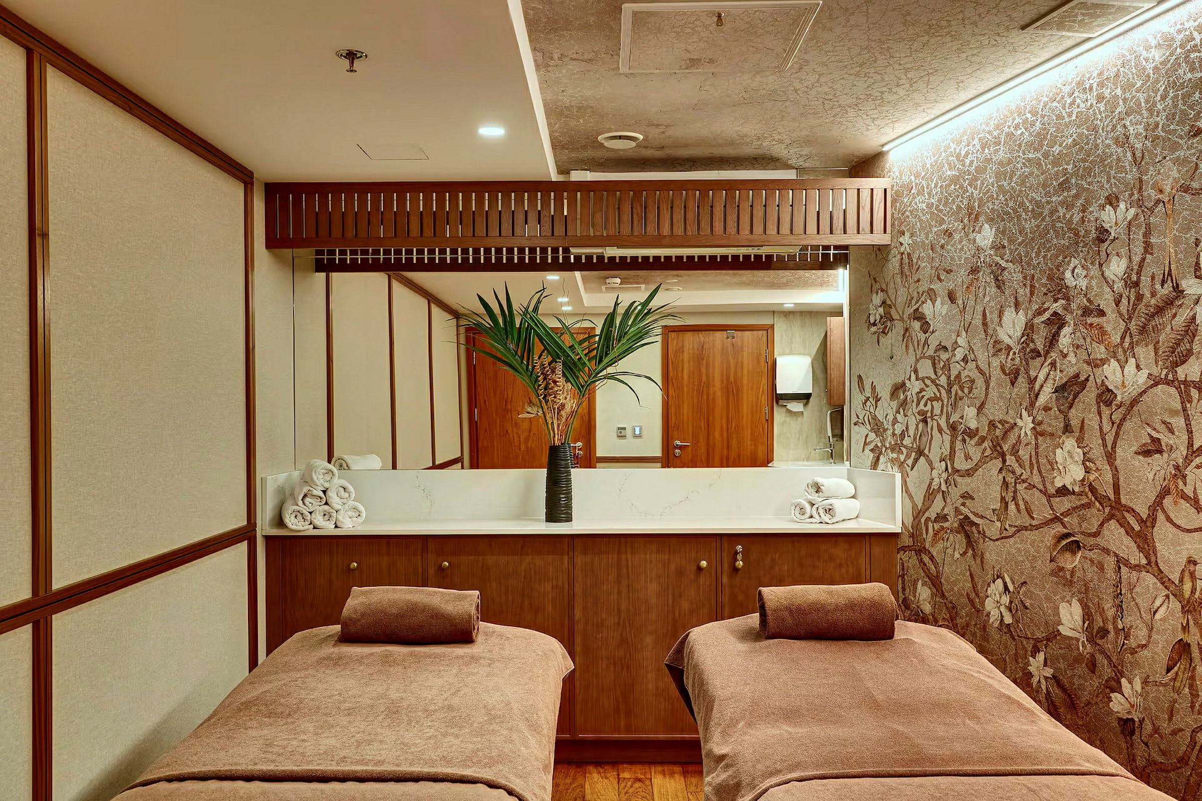 Ett spa-rum med två massagebäddar täckta med bruna handdukar, en bänk med handdukar och en vas med palmblad framför en spegel. (AI-genererad text)
