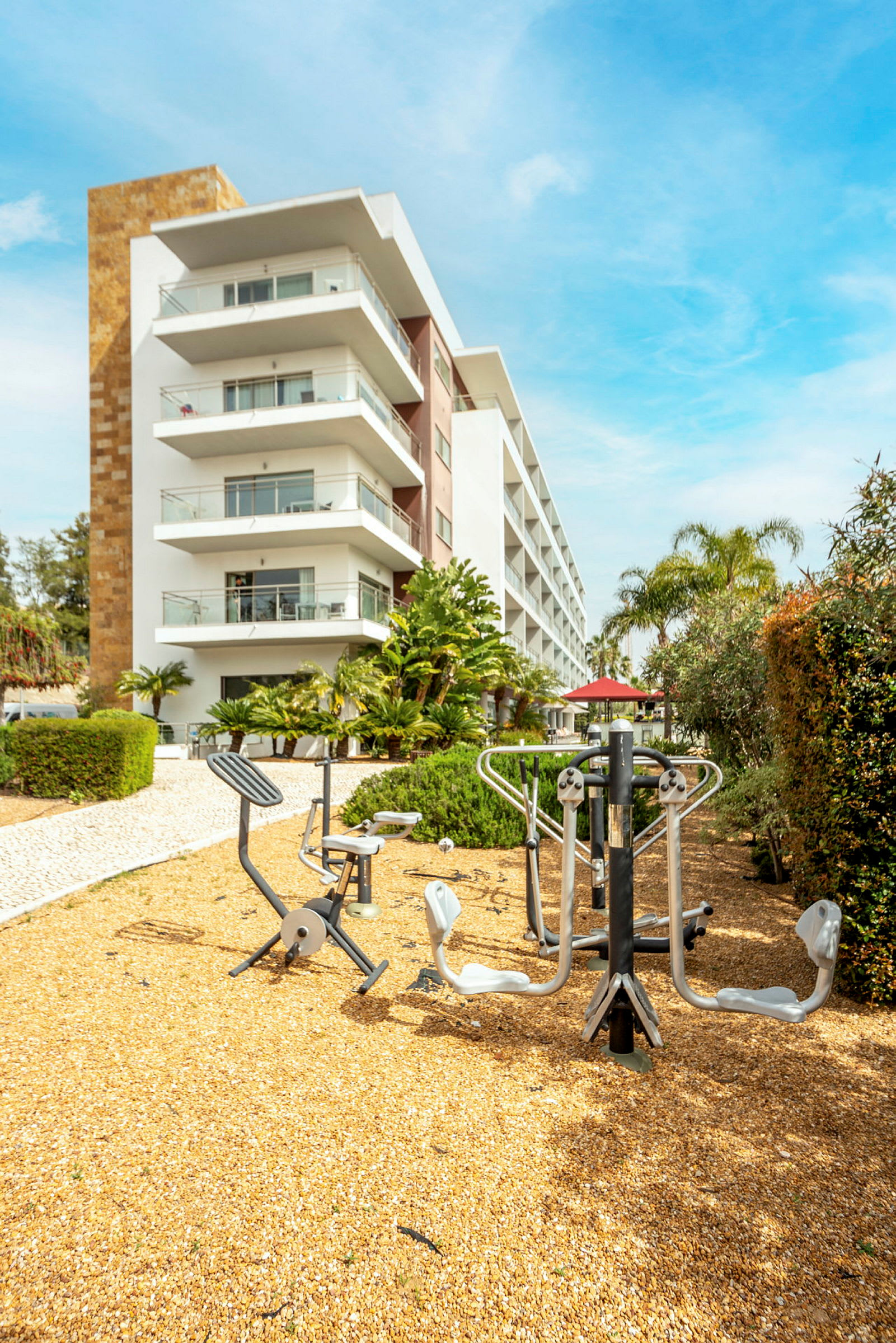 Utegym med träningsredskap framför en modern byggnad med flera våningar och balkonger, omgiven av grönska. (AI-genererad text)