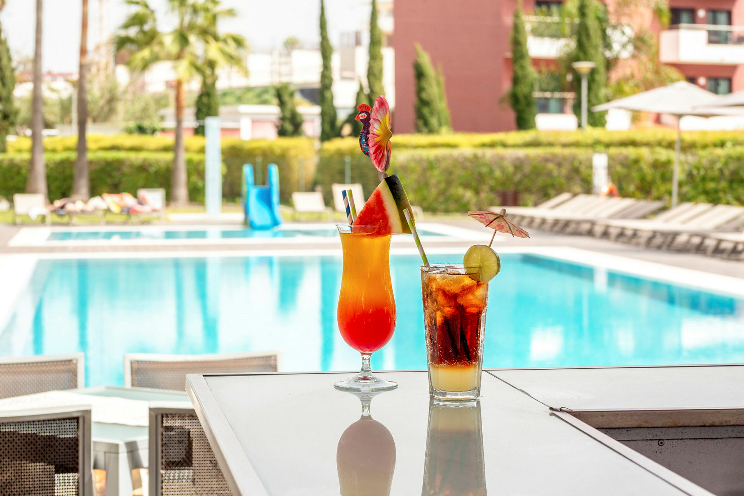 Två färgglada cocktails på ett bord med en swimmingpool i bakgrunden. (AI-genererad text)