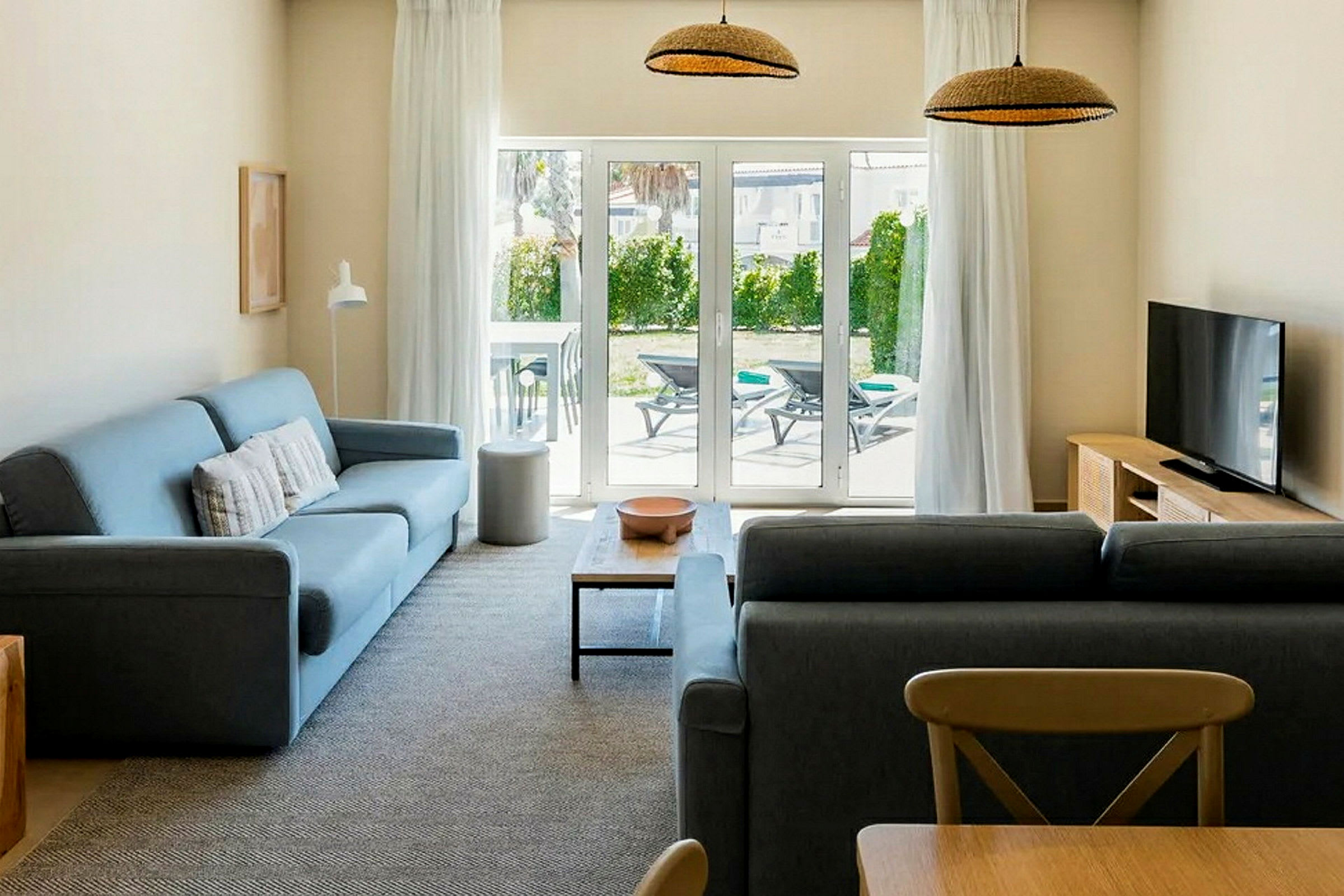 Ett vardagsrum med två soffor, ett soffbord, en TV och en glasdörr som vetter mot en uteplats med solstolar. (AI-genererad text)