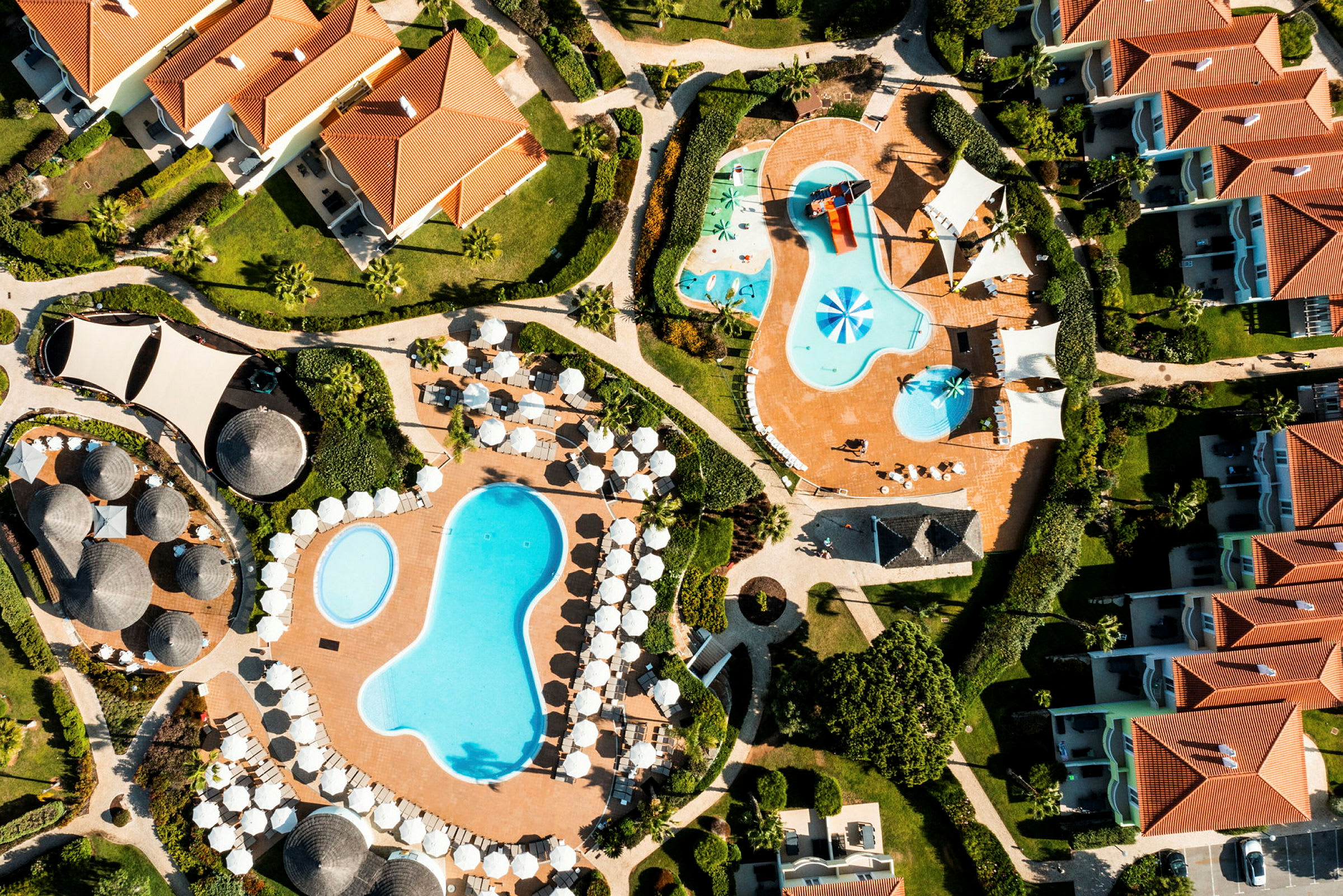 Flygvy över resortområde med flera pooler, solstolar, parasoller och byggnader. (AI-genererad text)