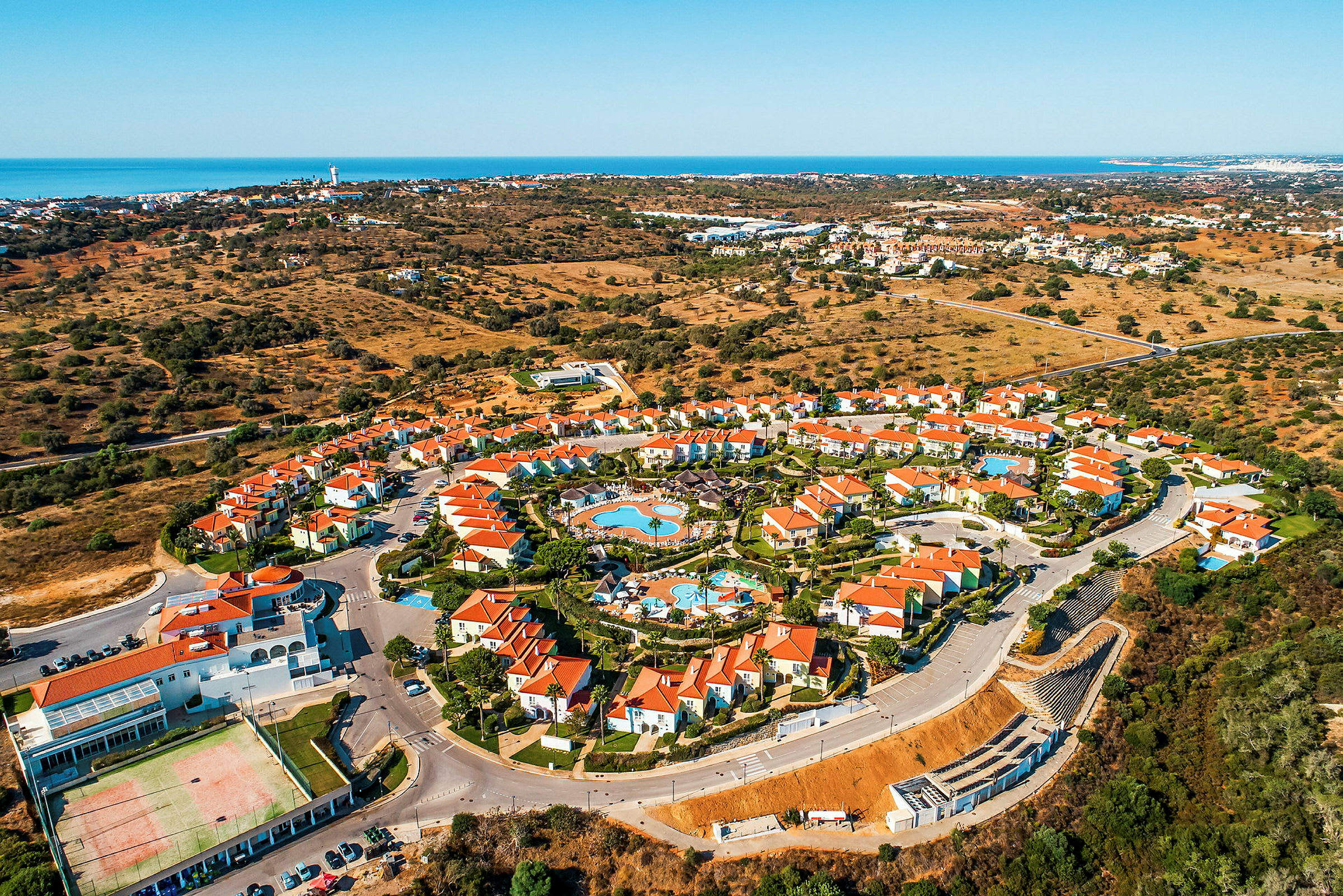 Flygfoto av en semesteranläggning med röda tak, omgiven av landskap och havet i bakgrunden. (AI-genererad text)