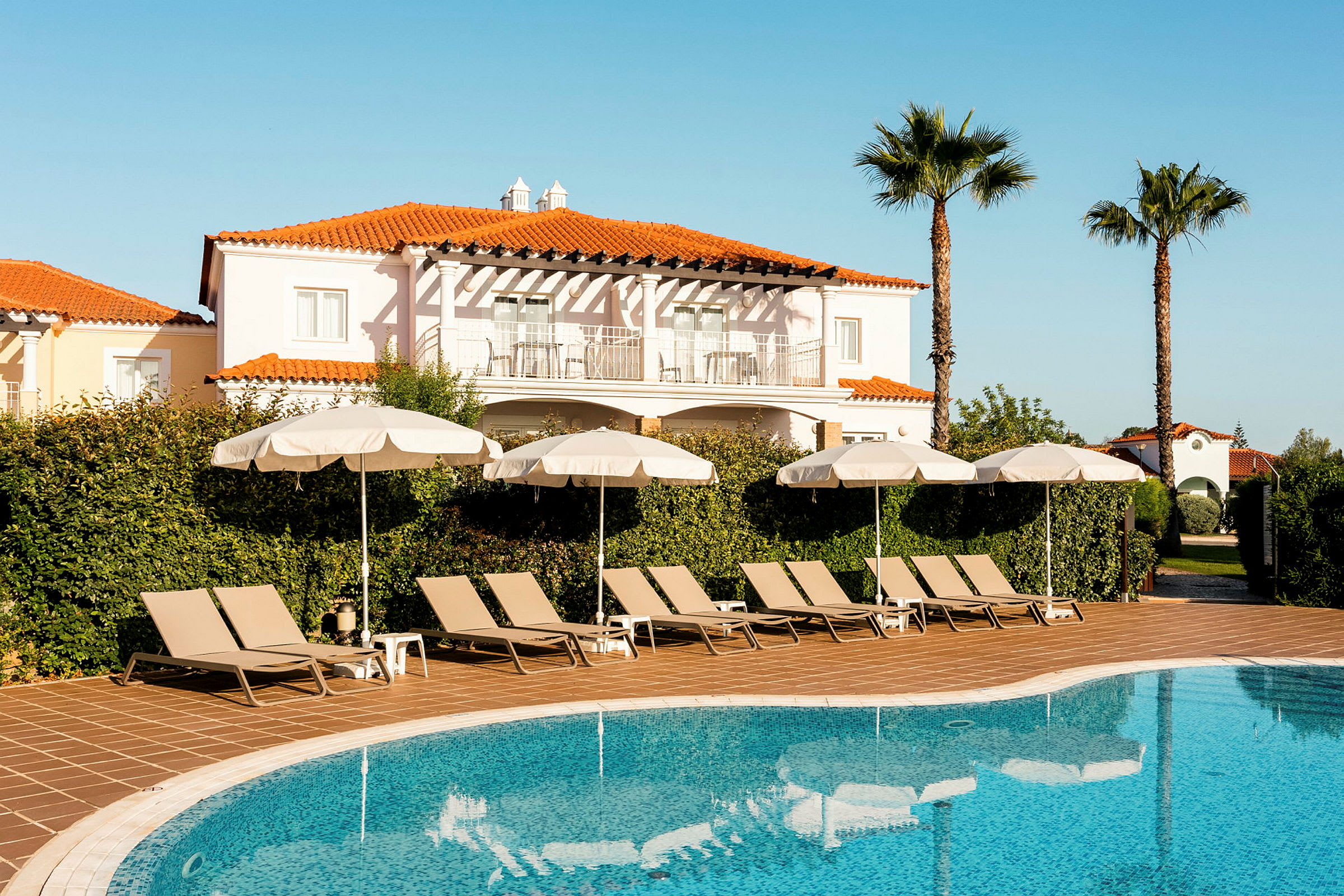 Pool med solstolar och parasoll framför en byggnad. (AI-genererad text)