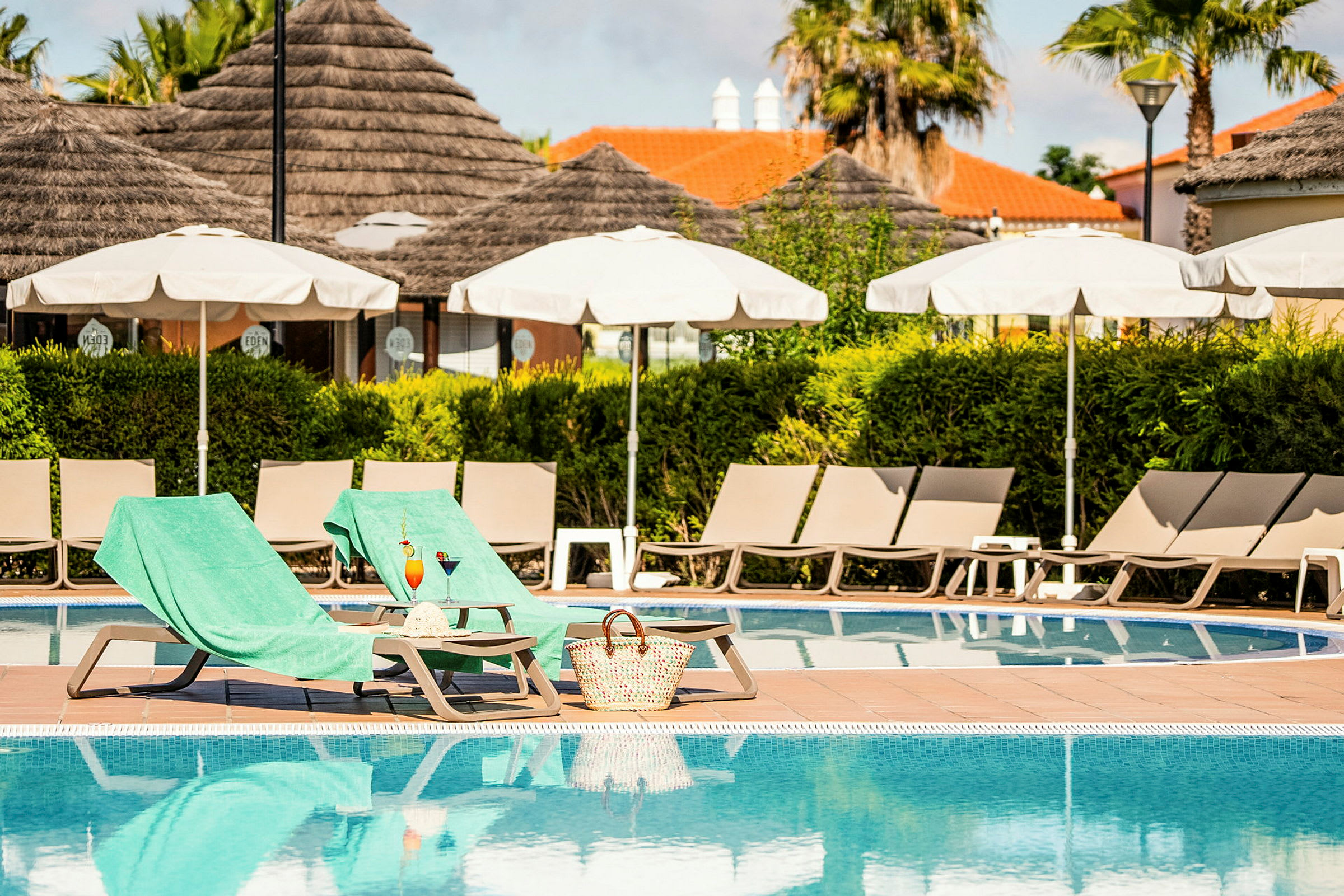 Två solstolar med gröna handdukar vid en pool, parasoller och växtlighet i bakgrunden. (AI-genererad text)