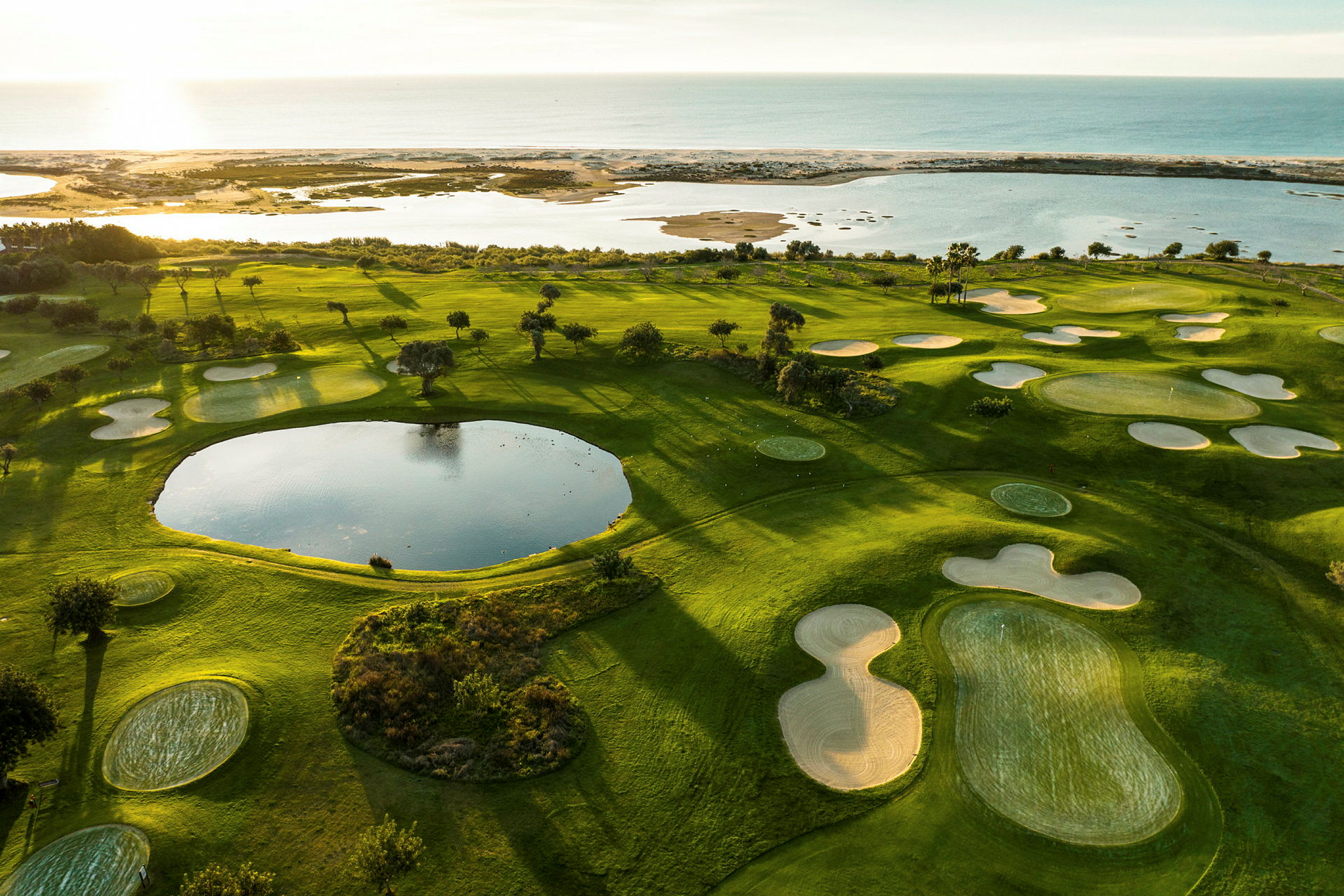 Vy över en grön golfbana med sandbunkrar och en damm, i närheten av kust och hav. (AI-genererad text)
