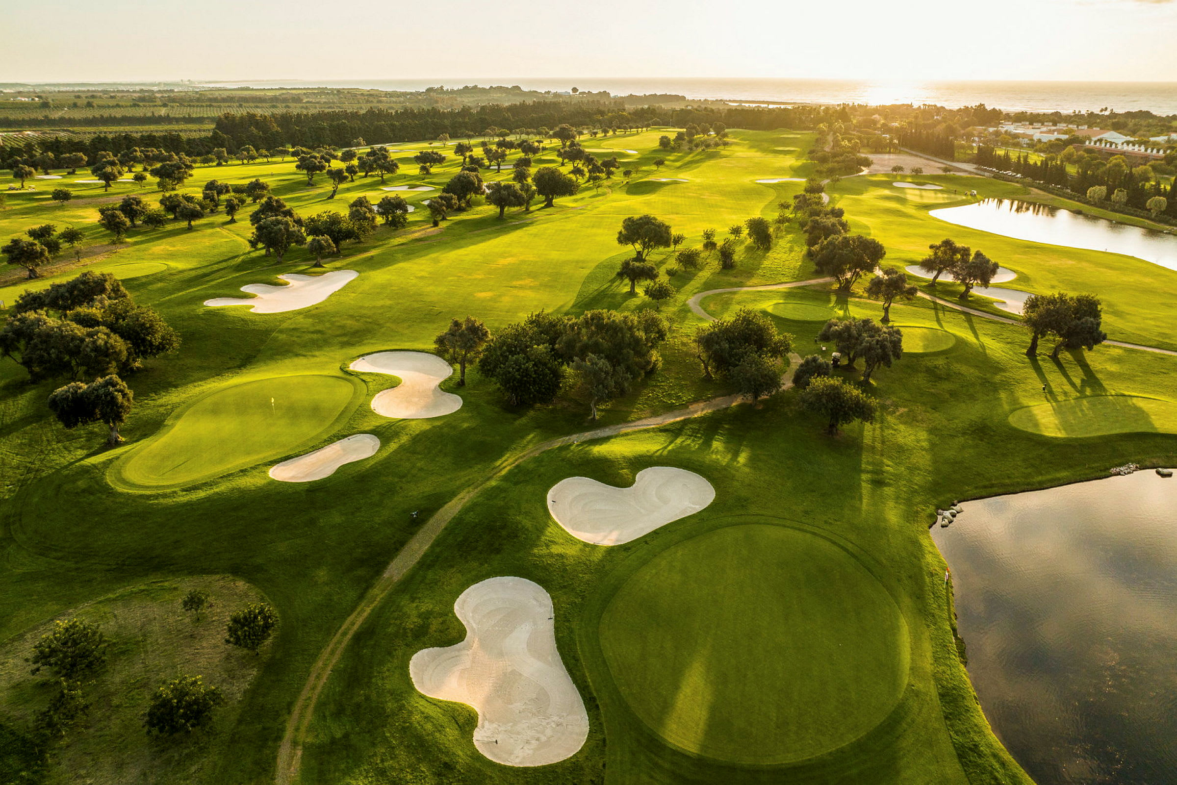 Flygvy över en golfbana med gröna fairways, sandbunkrar och vattenhinder vid solnedgången. (AI-genererad text)