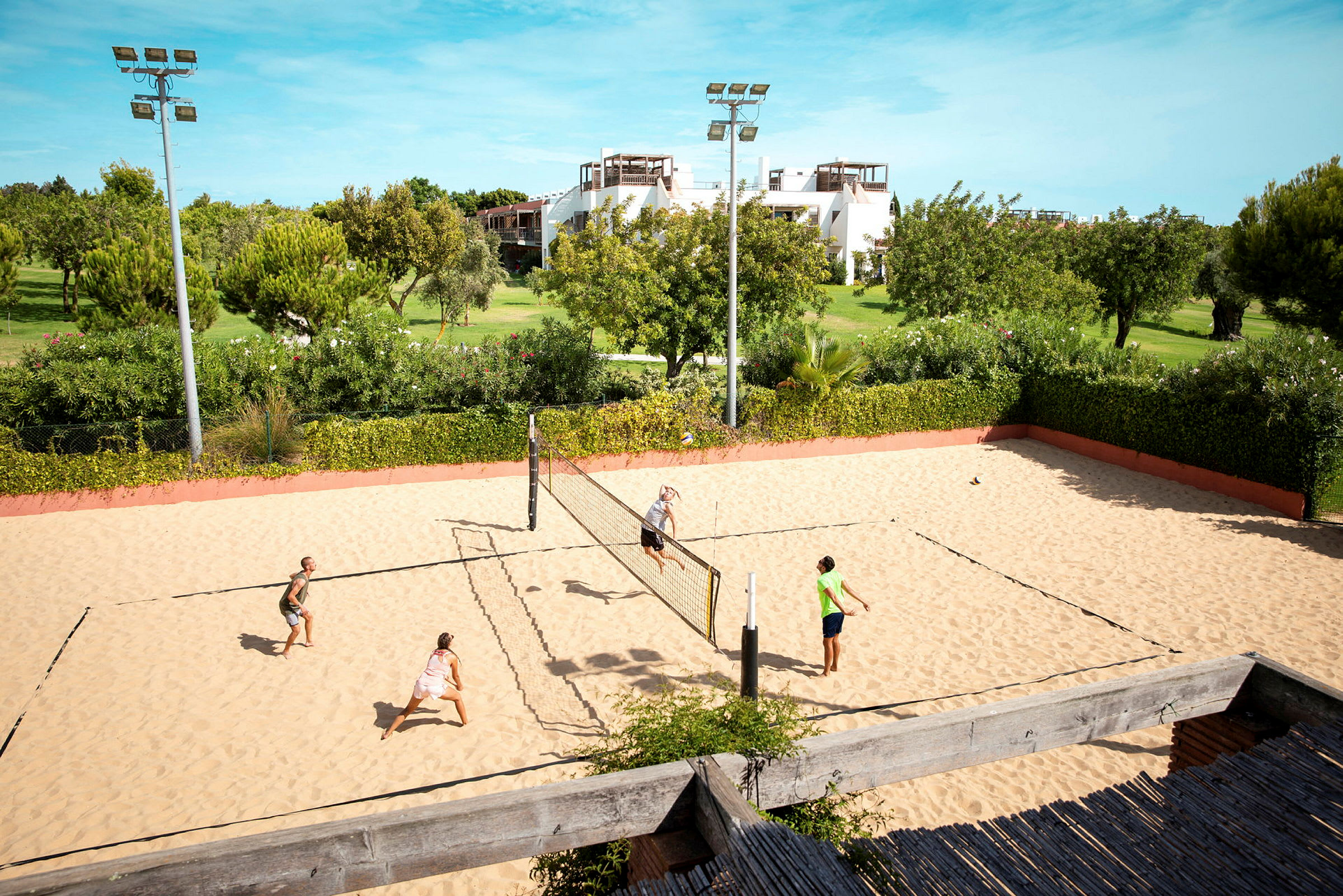 Sandvolleybollplan med fyra personer som spelar, omgiven av grönt landskap och byggnader i bakgrunden. (AI-genererad text)
