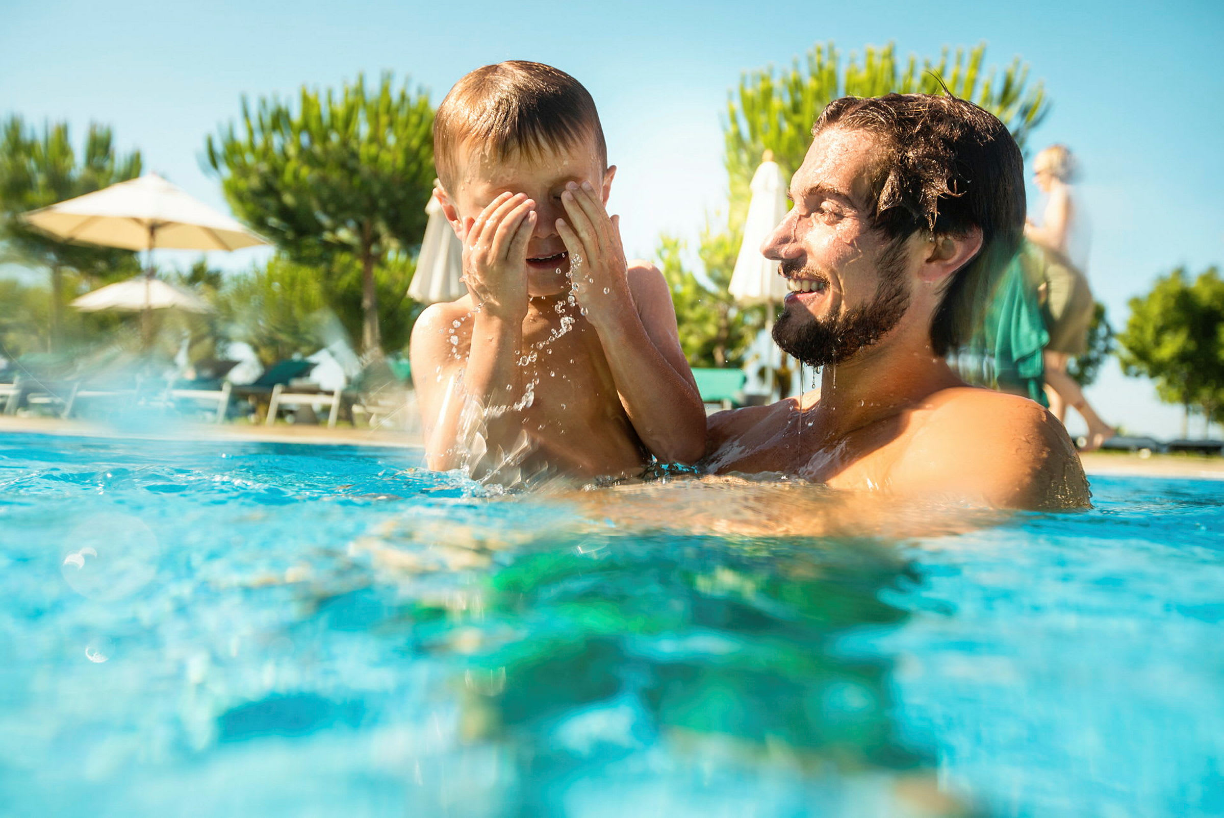 Personer njuter av en solig dag i en pool med parasoller och träd i bakgrunden. (AI-genererad text)