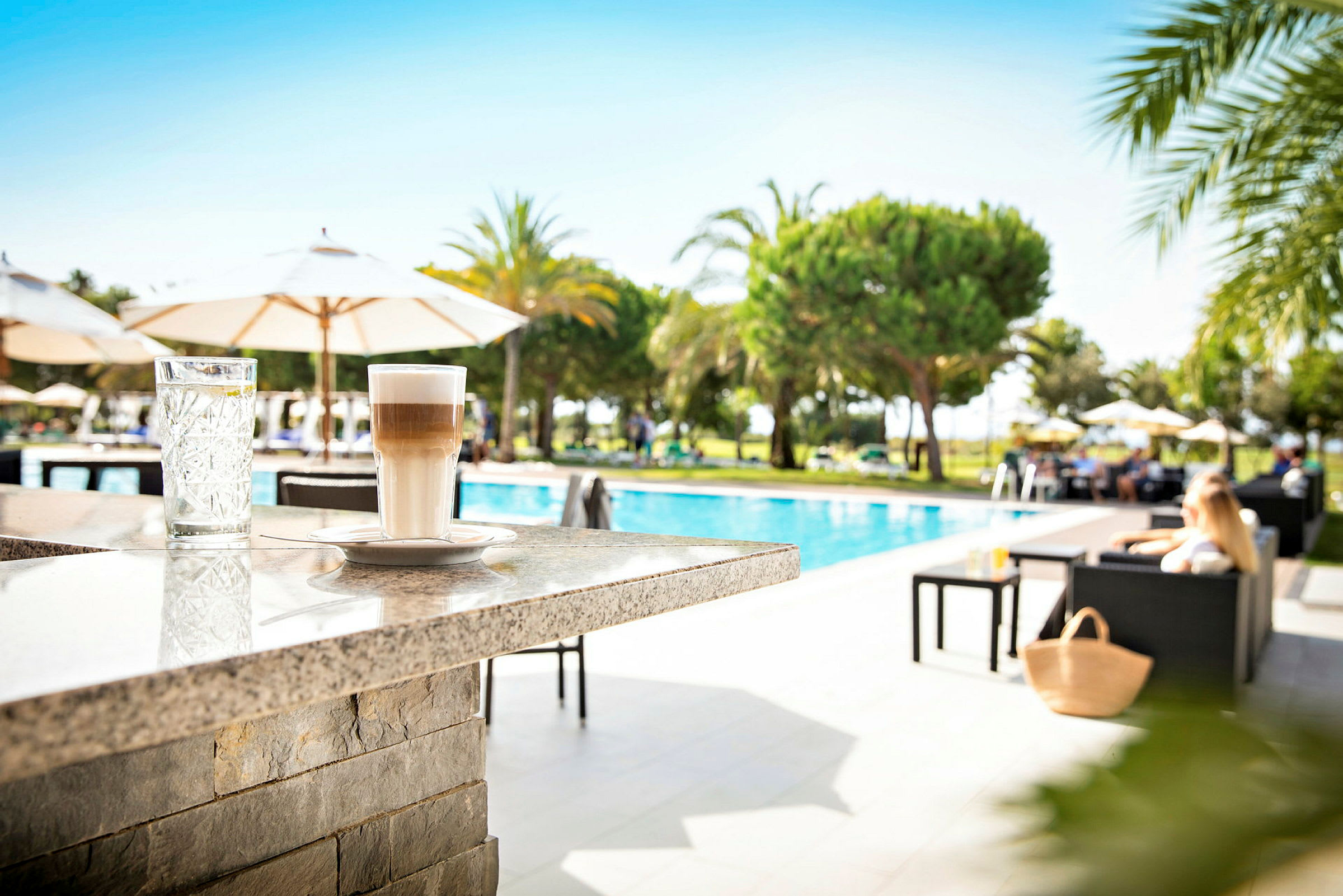 Ett glas vatten och en latte på en stenbar vid en pool med omgivande solstolar och parasoller. (AI-genererad text)