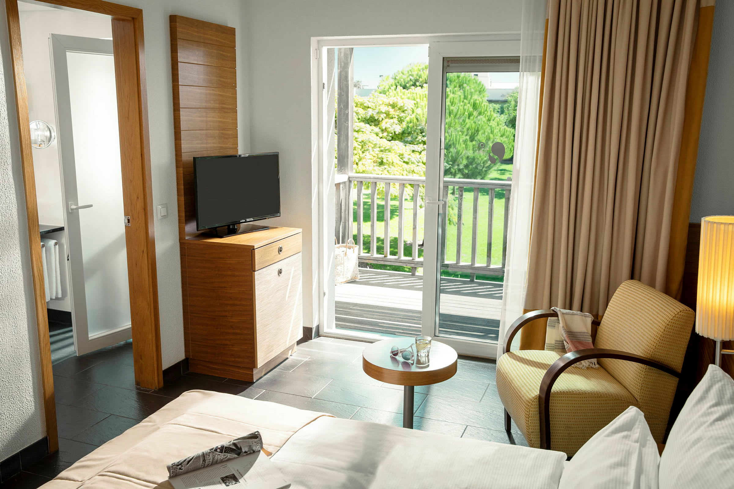 Ett hotellrum med säng, fåtölj, bord, TV och en balkong som vetter mot en grönskande trädgård. (AI-genererad text)