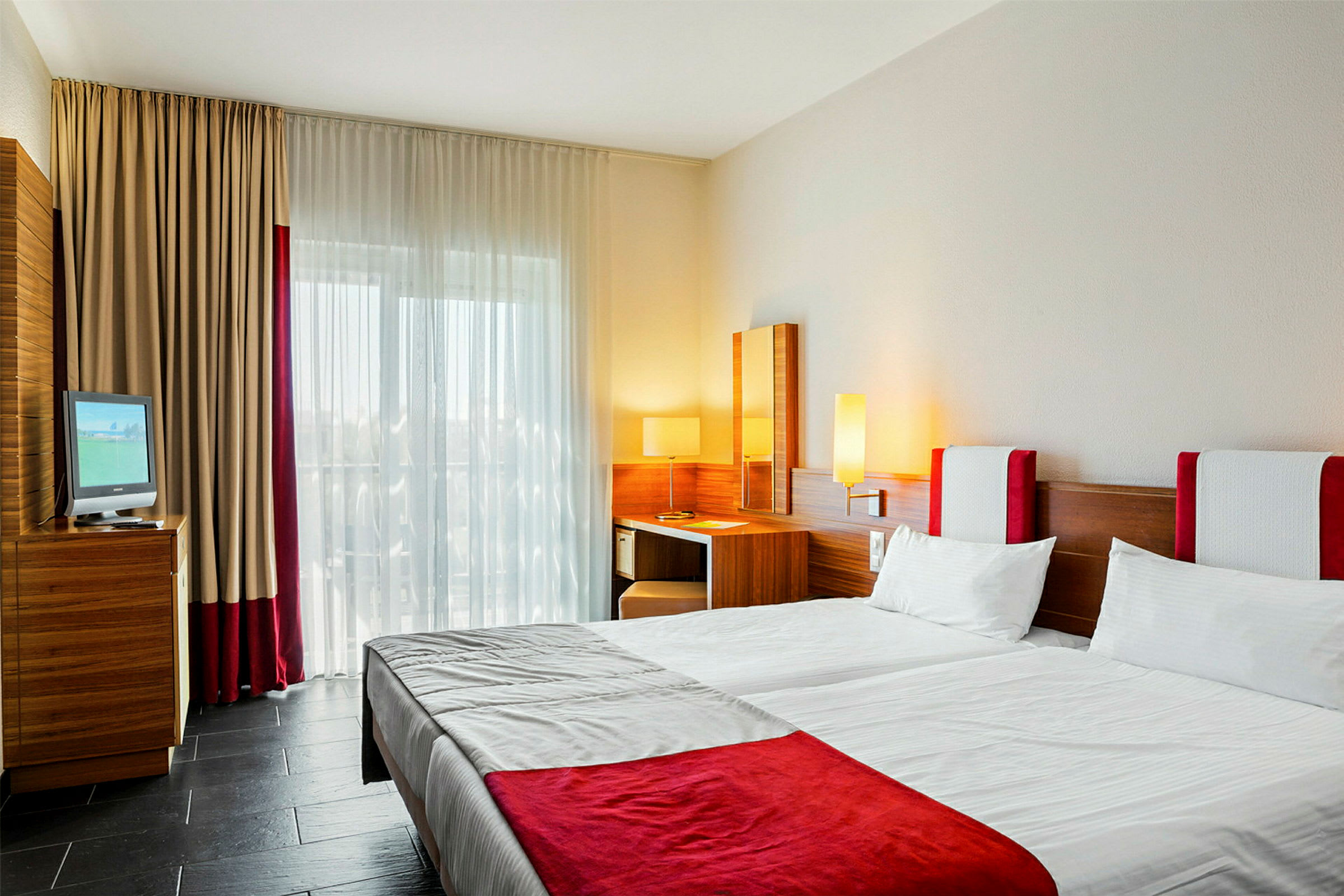 Hotellrum med två sängar, skrivbord och fönster med gardiner. (AI-genererad text)