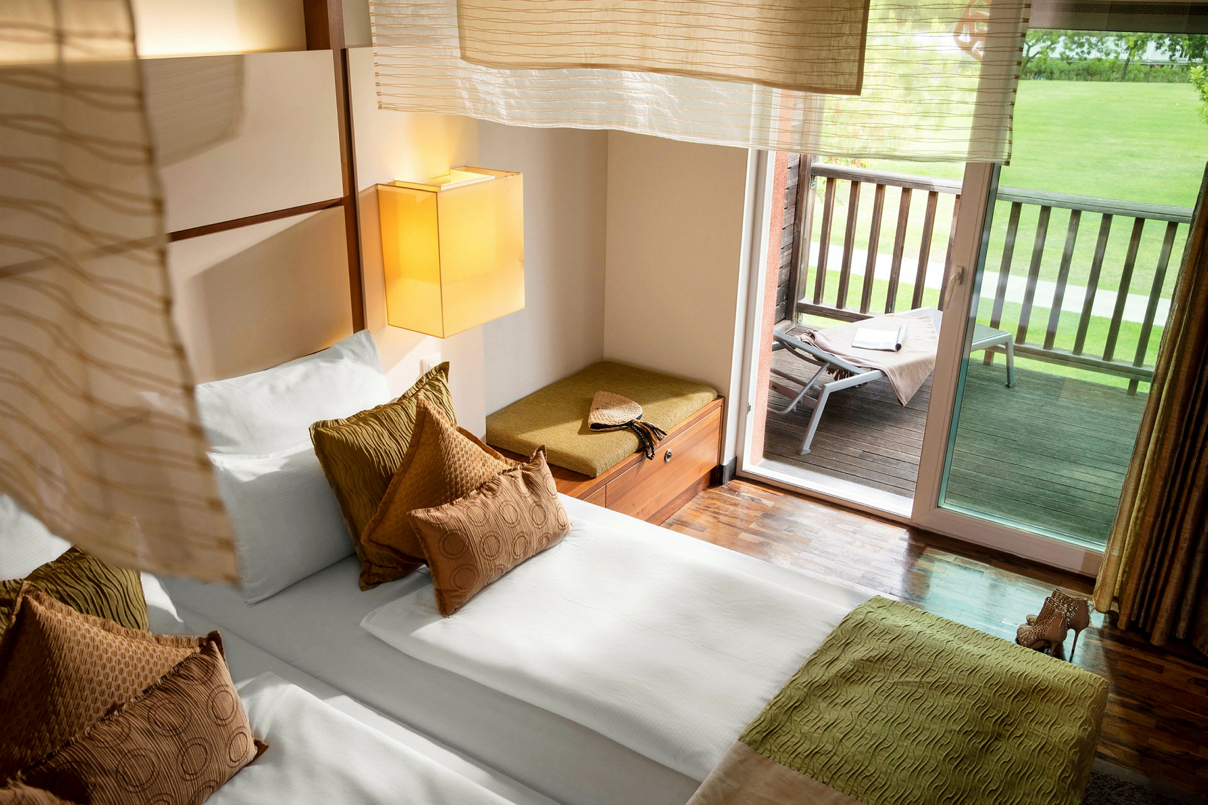Ett hotellrum med en säng och kuddar, sittbänk vid fönstret och en balkong med grönska. (AI-genererad text)