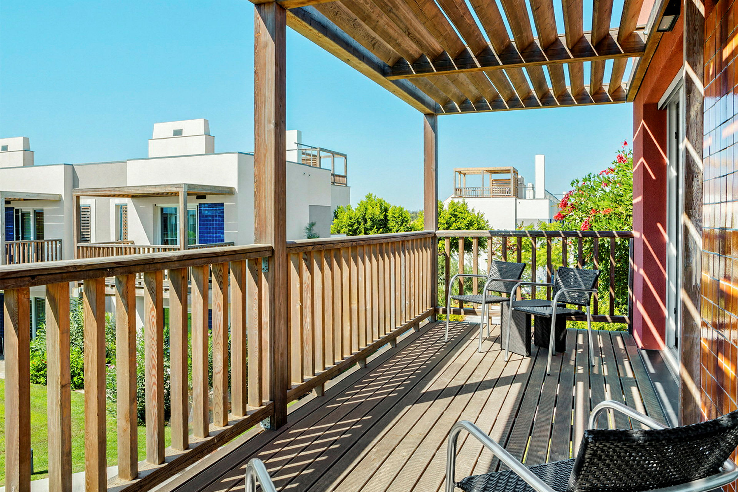 En balkong med trästaket och pergola med utsikt över andra byggnader och grönska. Stolar och ett litet bord står på balkongen. (AI-genererad text)