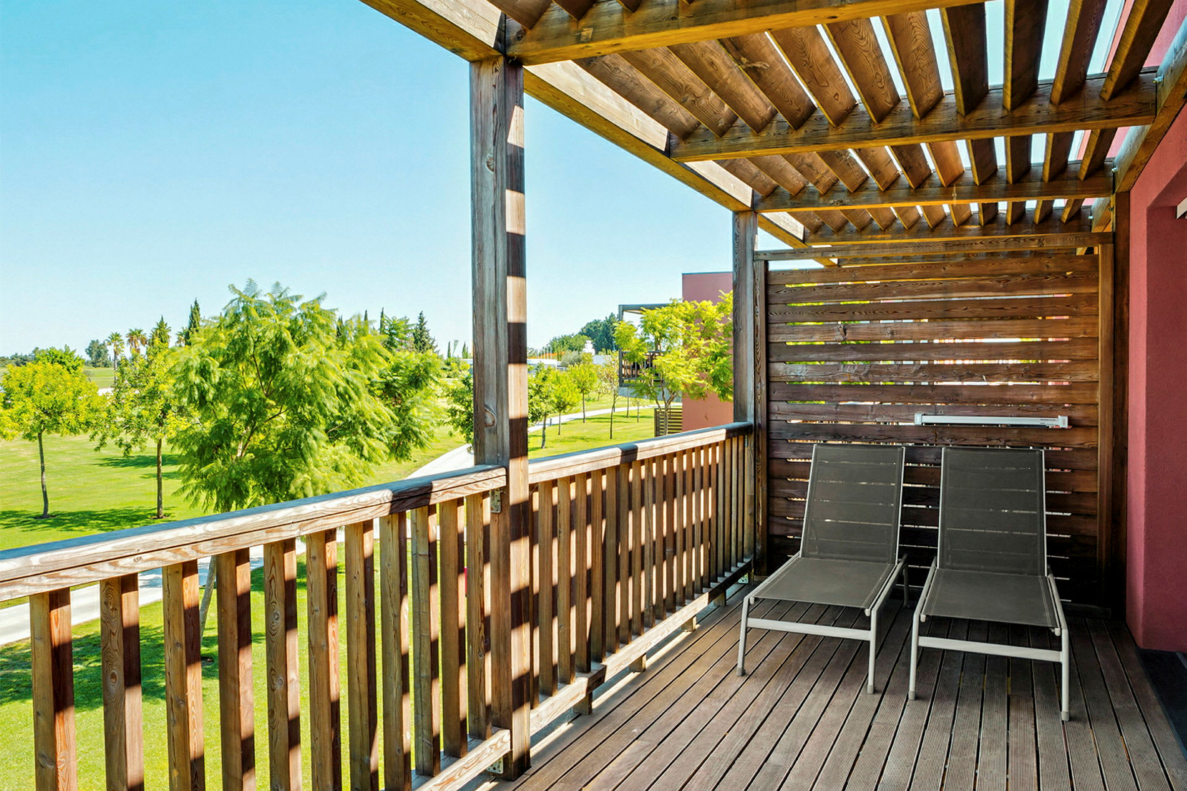 En träveranda med två solstolar stående under ett tak av trälameller, omgärdad av ett stängsel, med utsikt över ett grönt fält och några träd. (AI-genererad text)