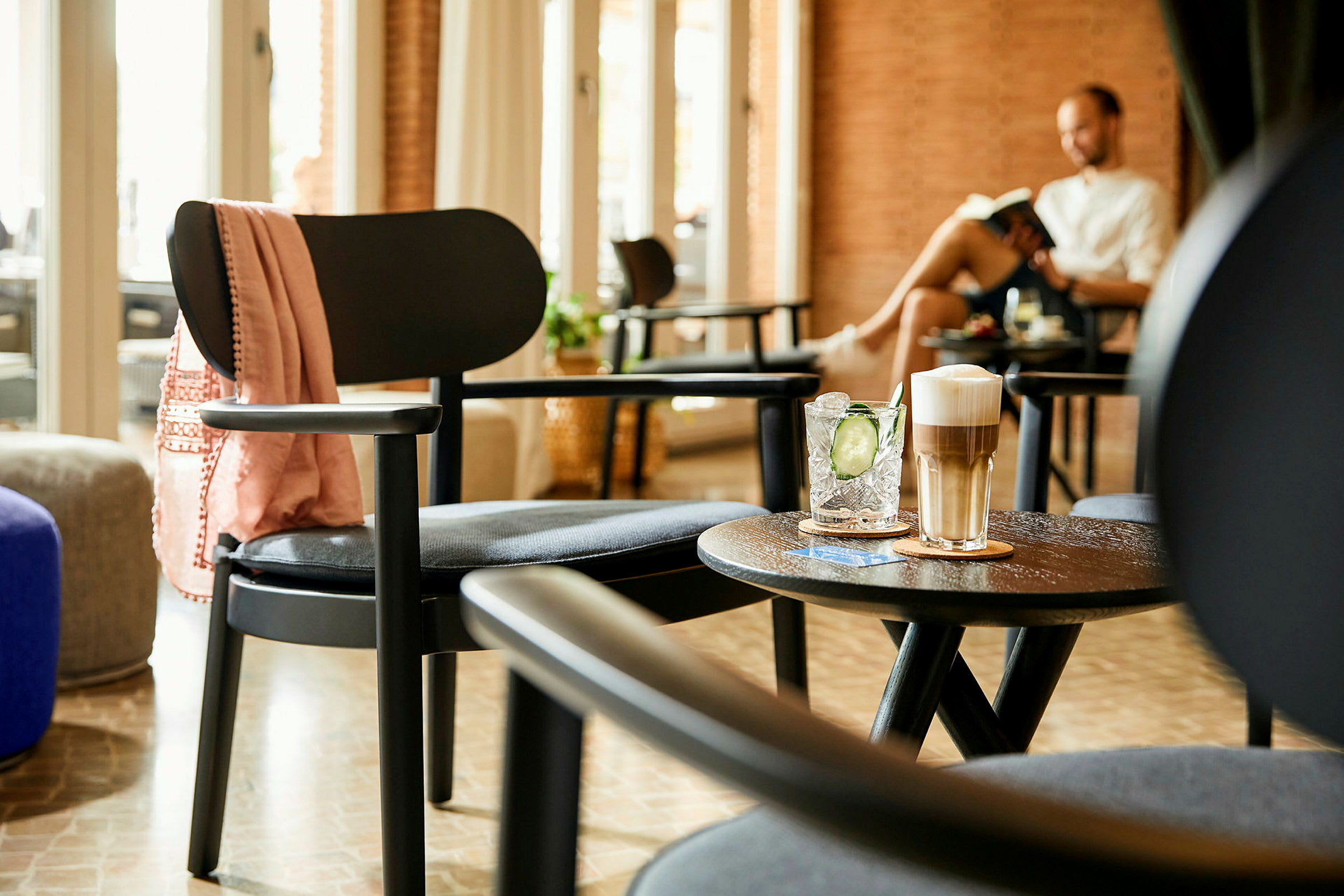 En interiörbild som visar en sittgrupp med stolar, bord och drycker, med en person som läser i bakgrunden. (AI-genererad text)