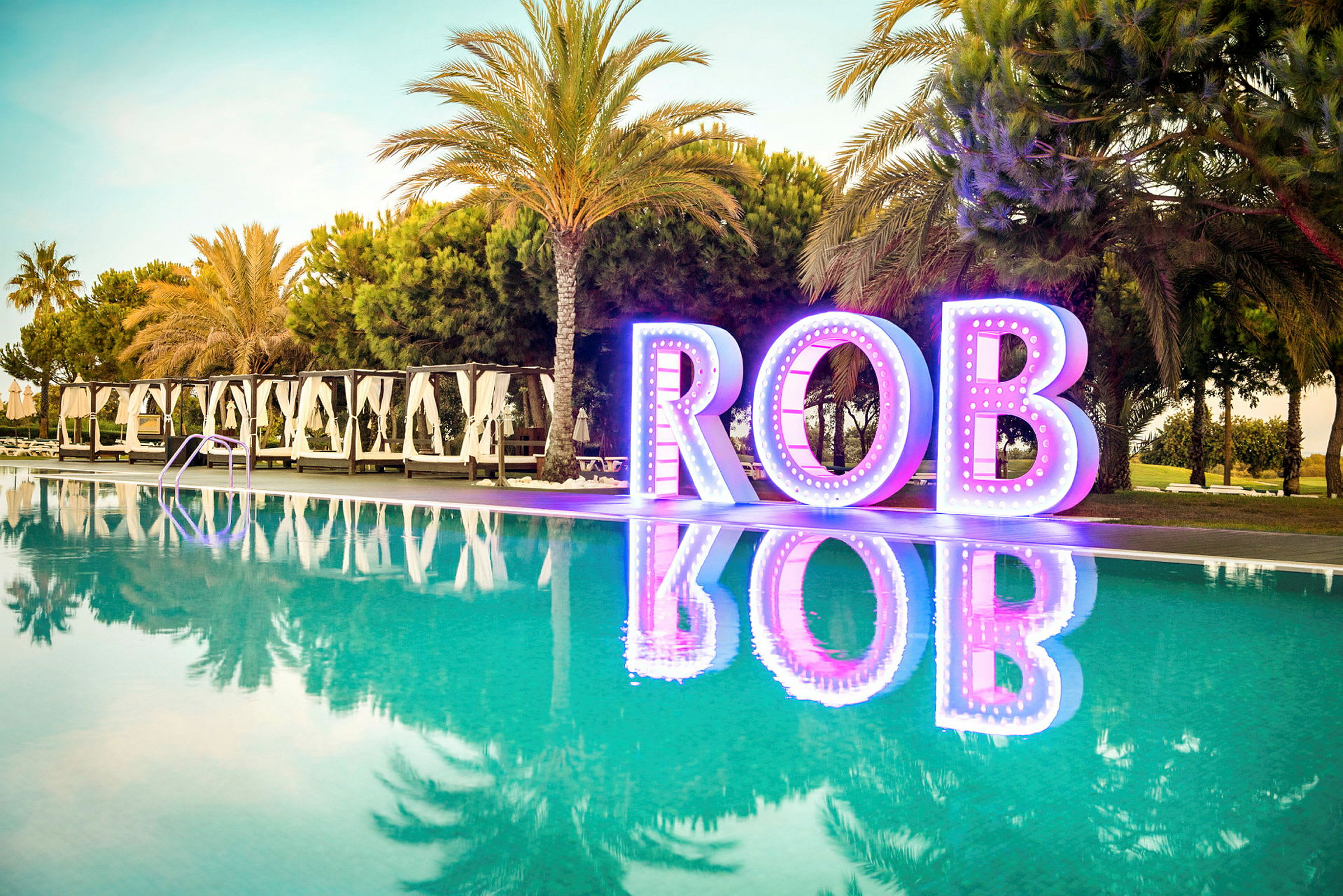 En pool med palmträd och vita cabanas i bakgrunden. Stora upplysta bokstäver som bildar ordet 'ROB' vid poolkanten. (AI-genererad text)