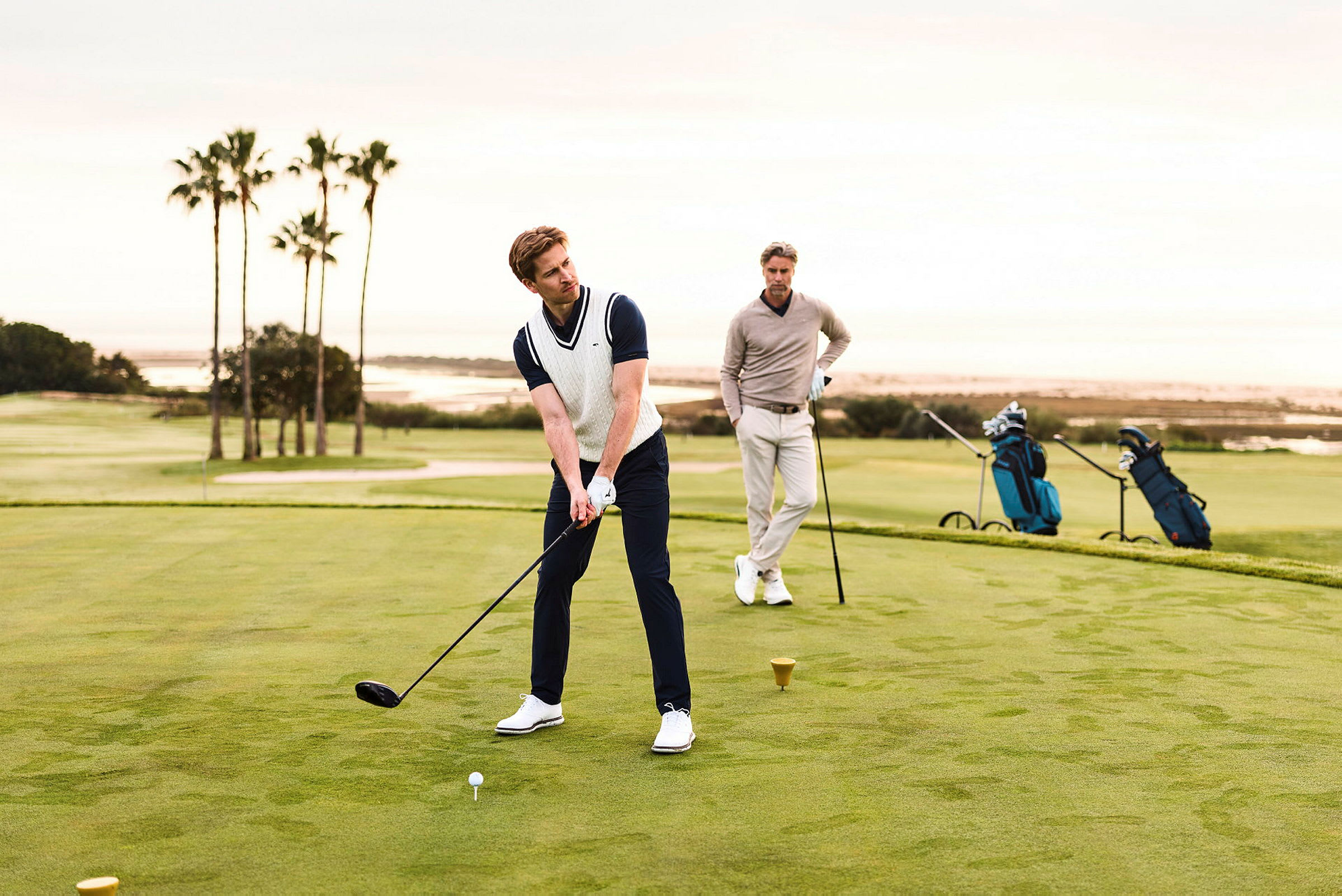 En person spelar golf och en annan står bredvid, med en grön golfbana i bakgrunden. (AI-genererad text)