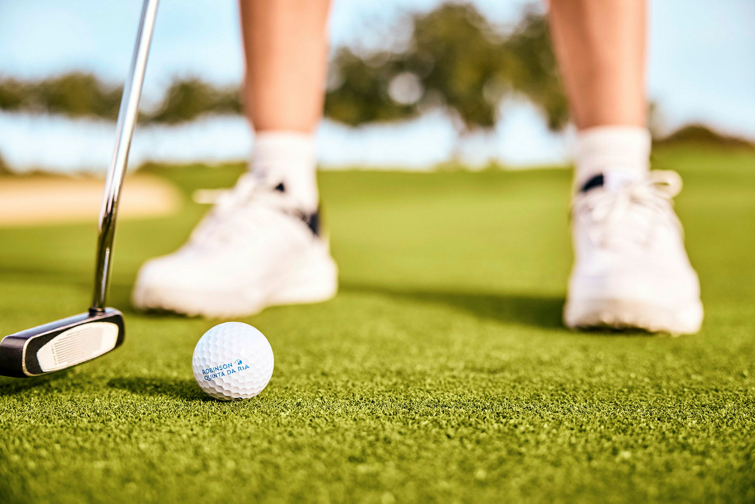 En golfboll och en putter på en grön gräsplan, med en persons fötter iförda vita sportskor i bakgrunden. (AI-genererad text)