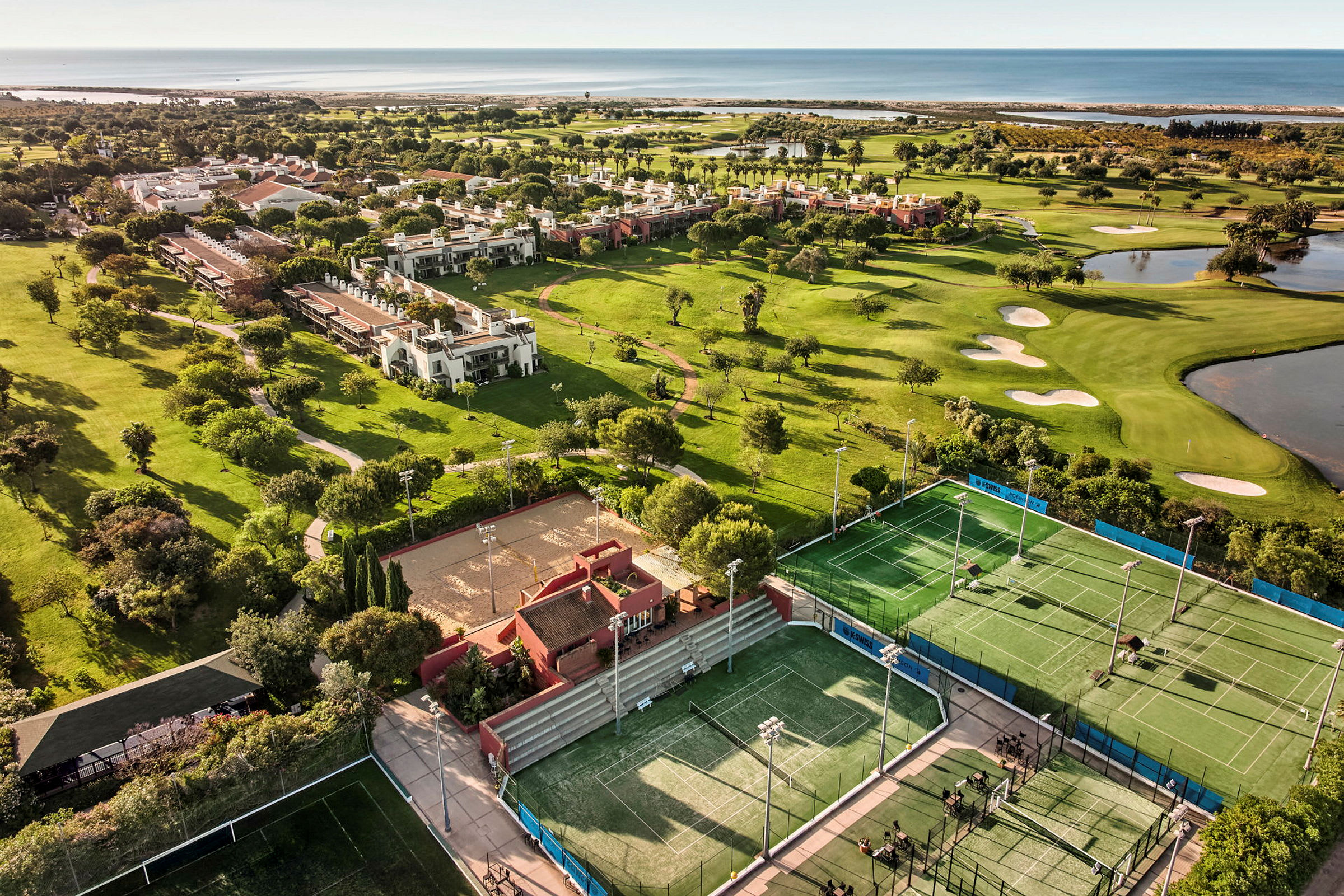 Flygfoto över en resort med tennisbanor, en golfbana och närliggande byggnader inbäddade i grönska och nära havet. (AI-genererad text)