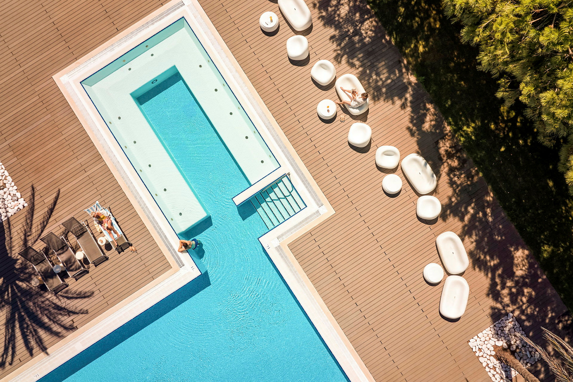 Flygbild av en pool omgiven av ett däck med loungestolar och sittmöbler, samt människor som kopplar av. (AI-genererad text)