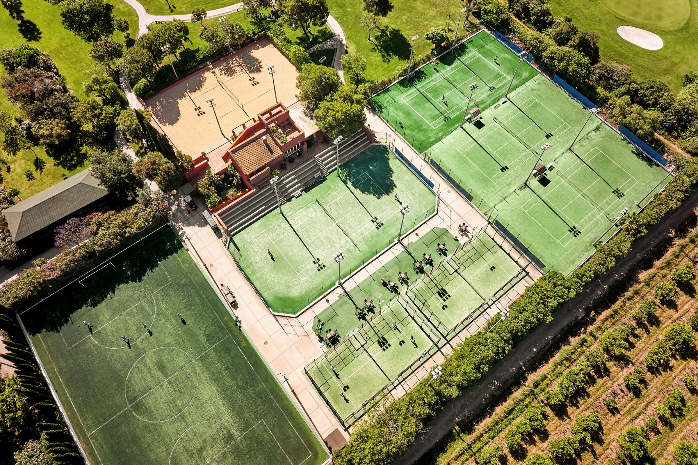 Flygvy över en sportanläggning med tennisbanor, padelbanor, en fotbollsplan och ett sandfält, omgiven av grönska. (AI-genererad text)