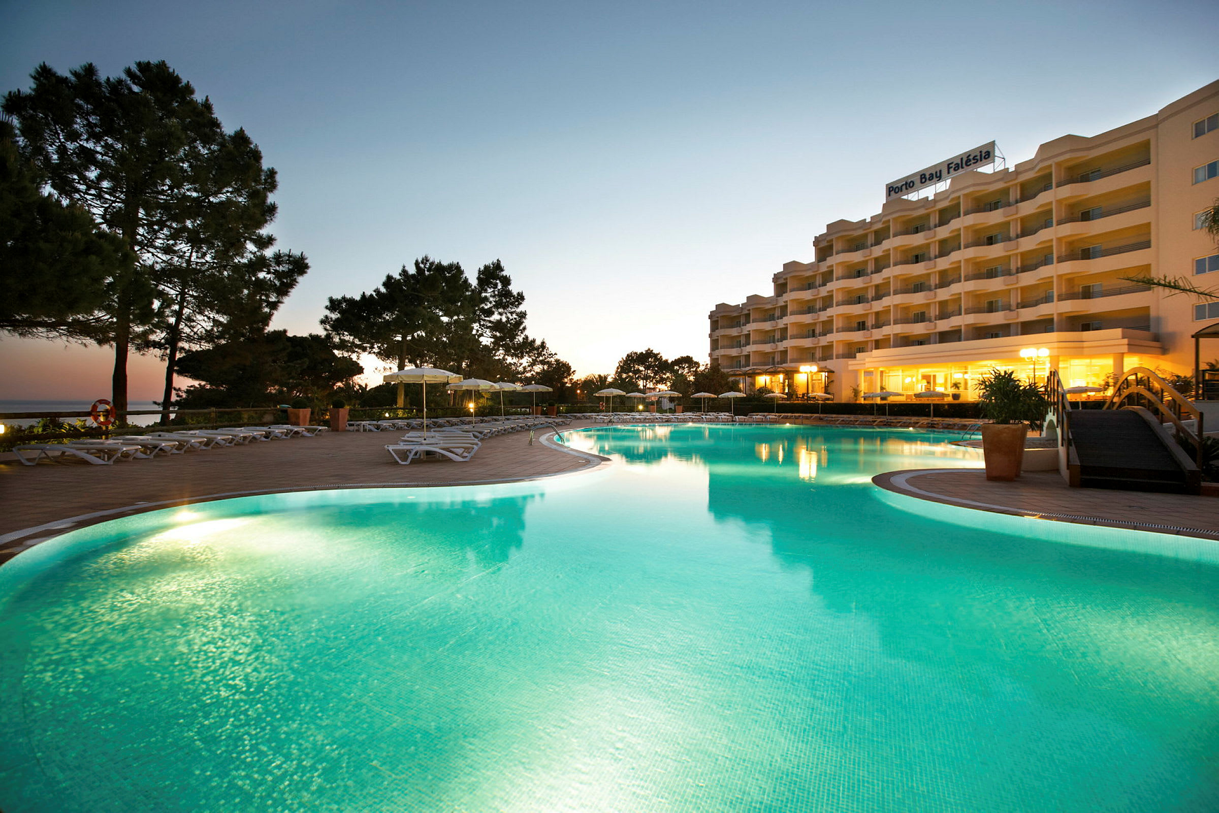 Bild på en hotellbyggnad med en stor pool i förgrunden, omgiven av solstolar och träd. Solnedgång syns i bakgrunden. (AI-genererad text)