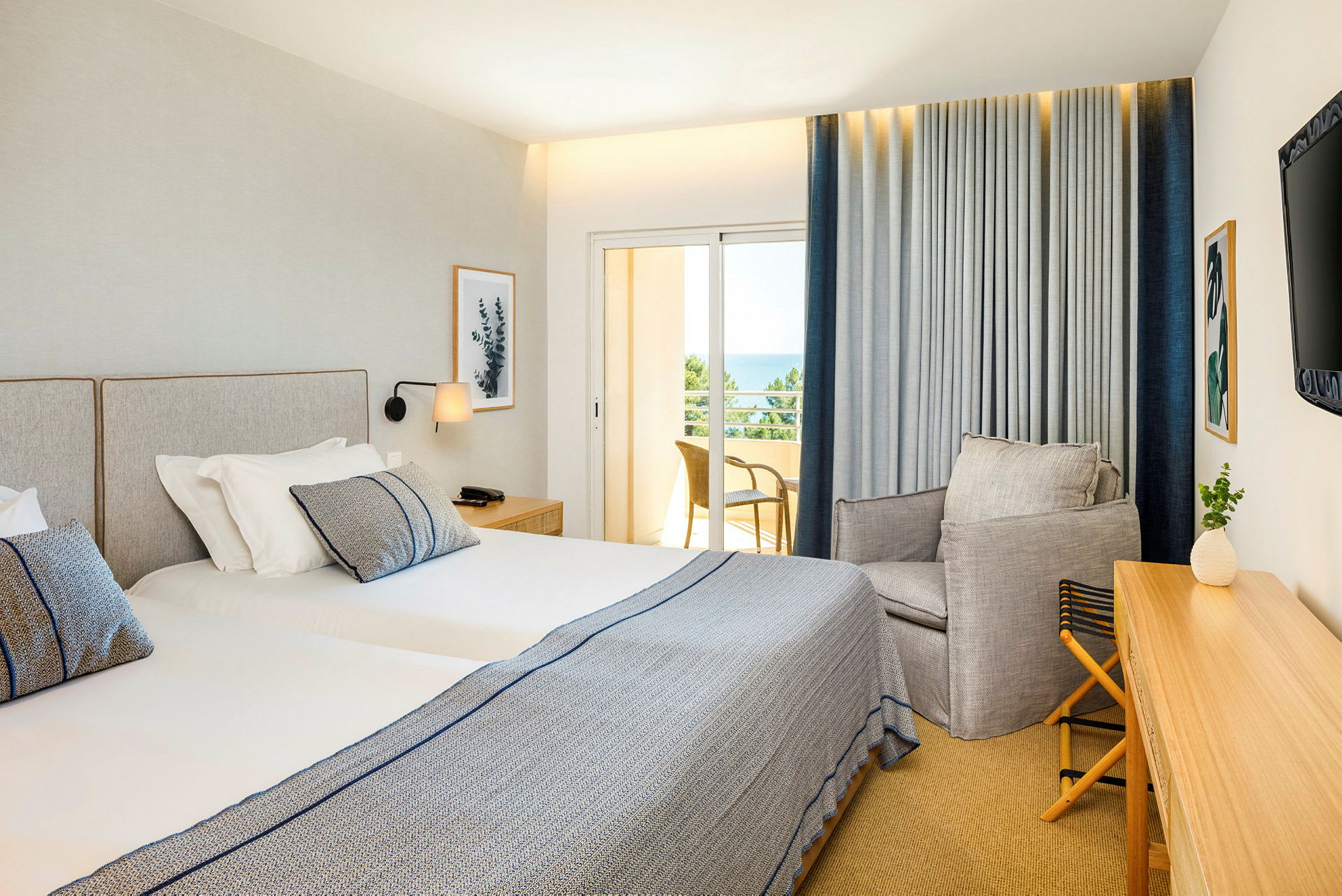 Ett hotellrum med två enkelsängar, en fåtölj och ett skrivbord. Balkongen har utsikt över havet. (AI-genererad text)