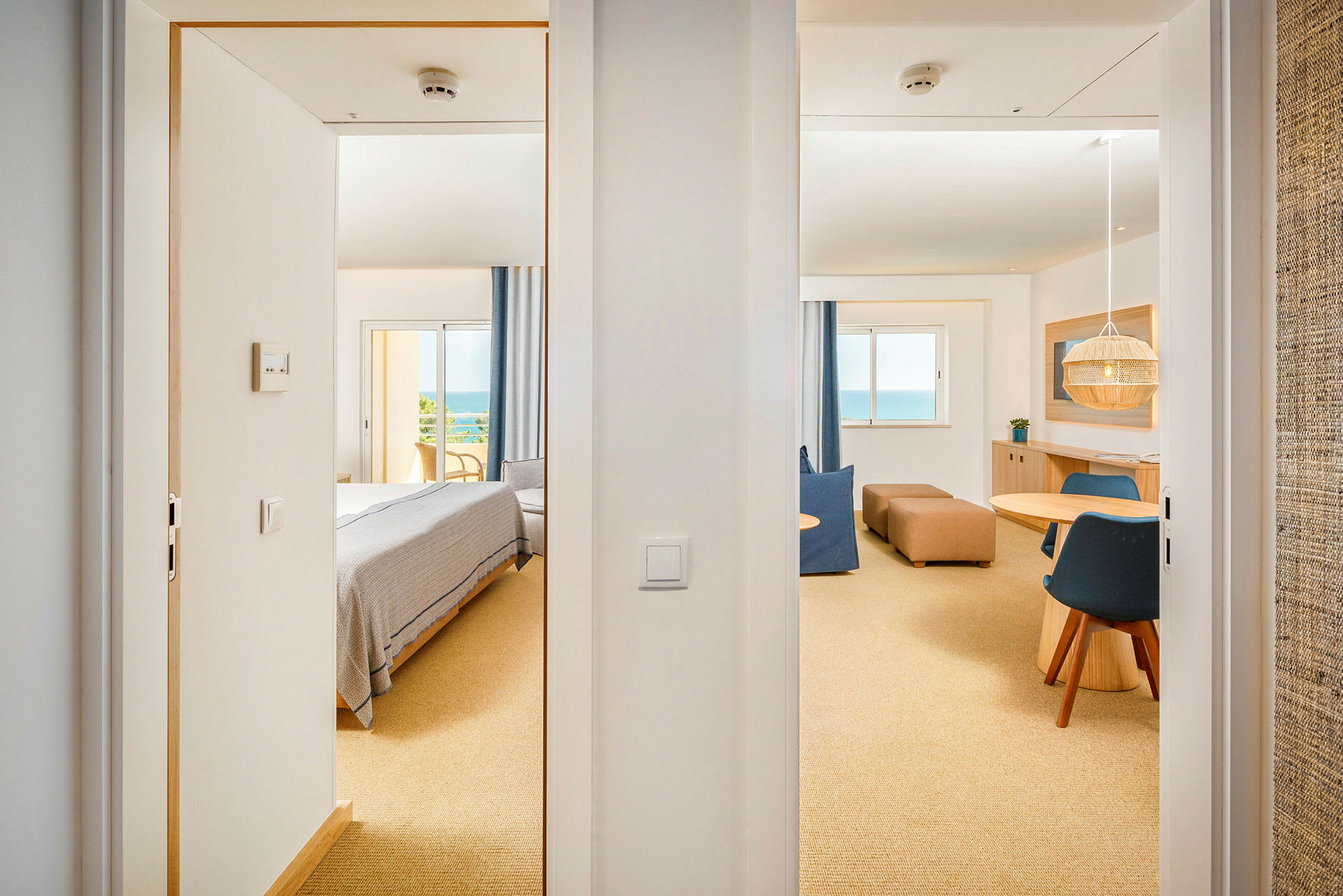 Utsikt genom två dörröppningar mot ett ljust hotellrum med sovdel och sittgrupp, samt ett stort fönster med havsutsikt. (AI-genererad text)