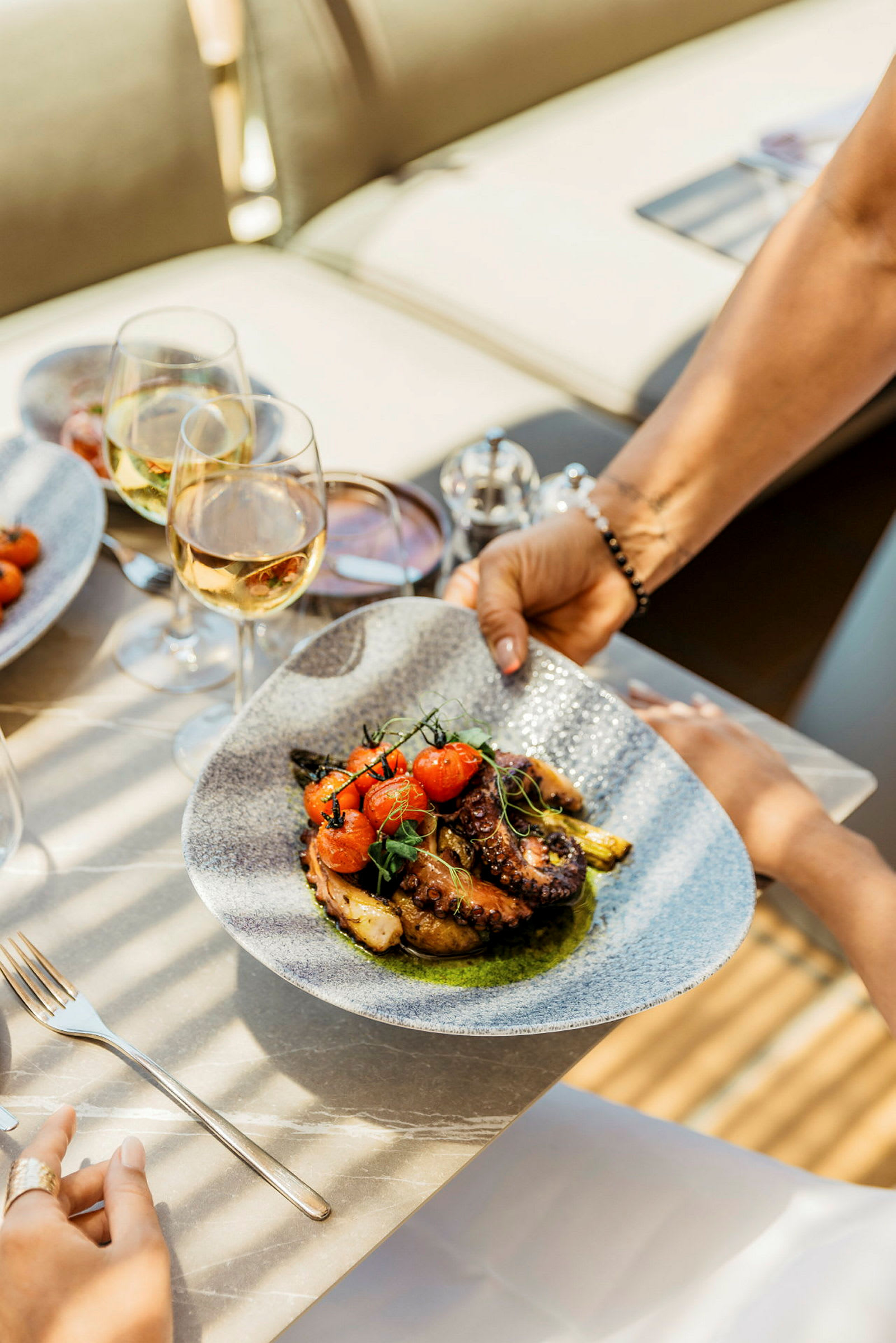 Bilden visar en tallrik med en delikat maträtt bestående av grillad bläckfisk, tomater och grönsaker, serverad på ett bord med glas vin. (AI-genererad text)