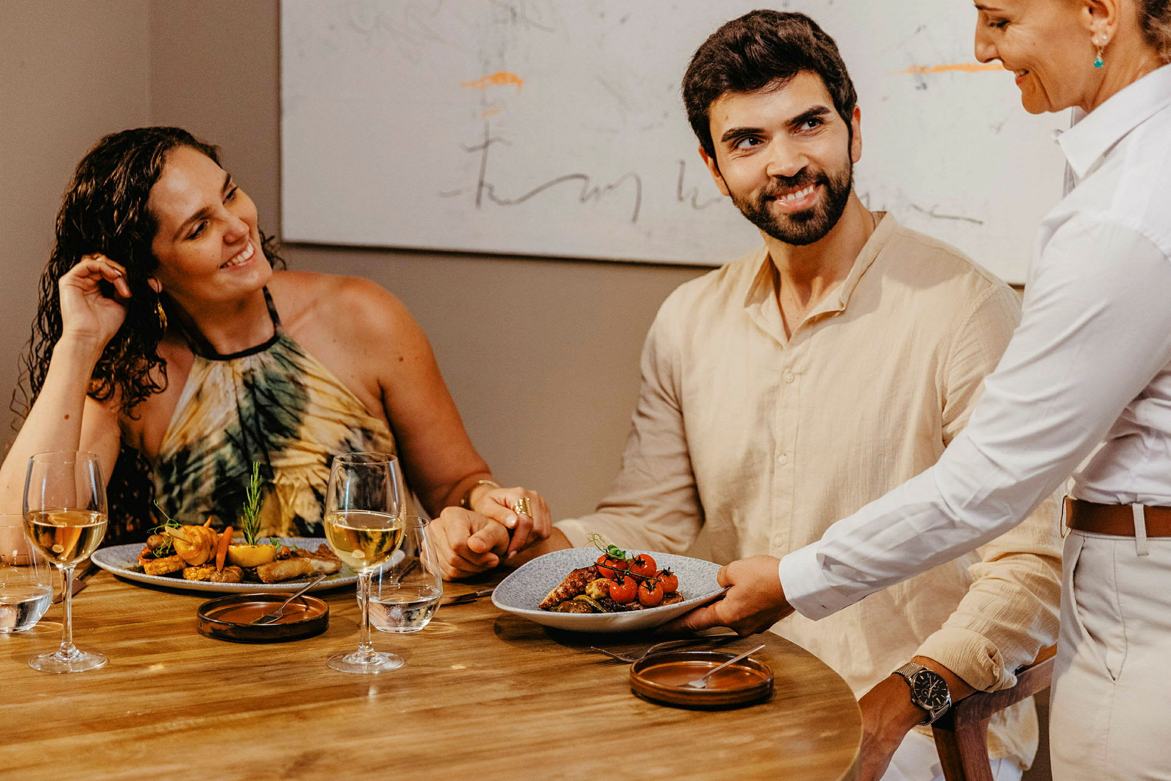 En servitör serverar en tallrik mat på ett bord där två personer sitter och håller varandras händer. (AI-genererad text)