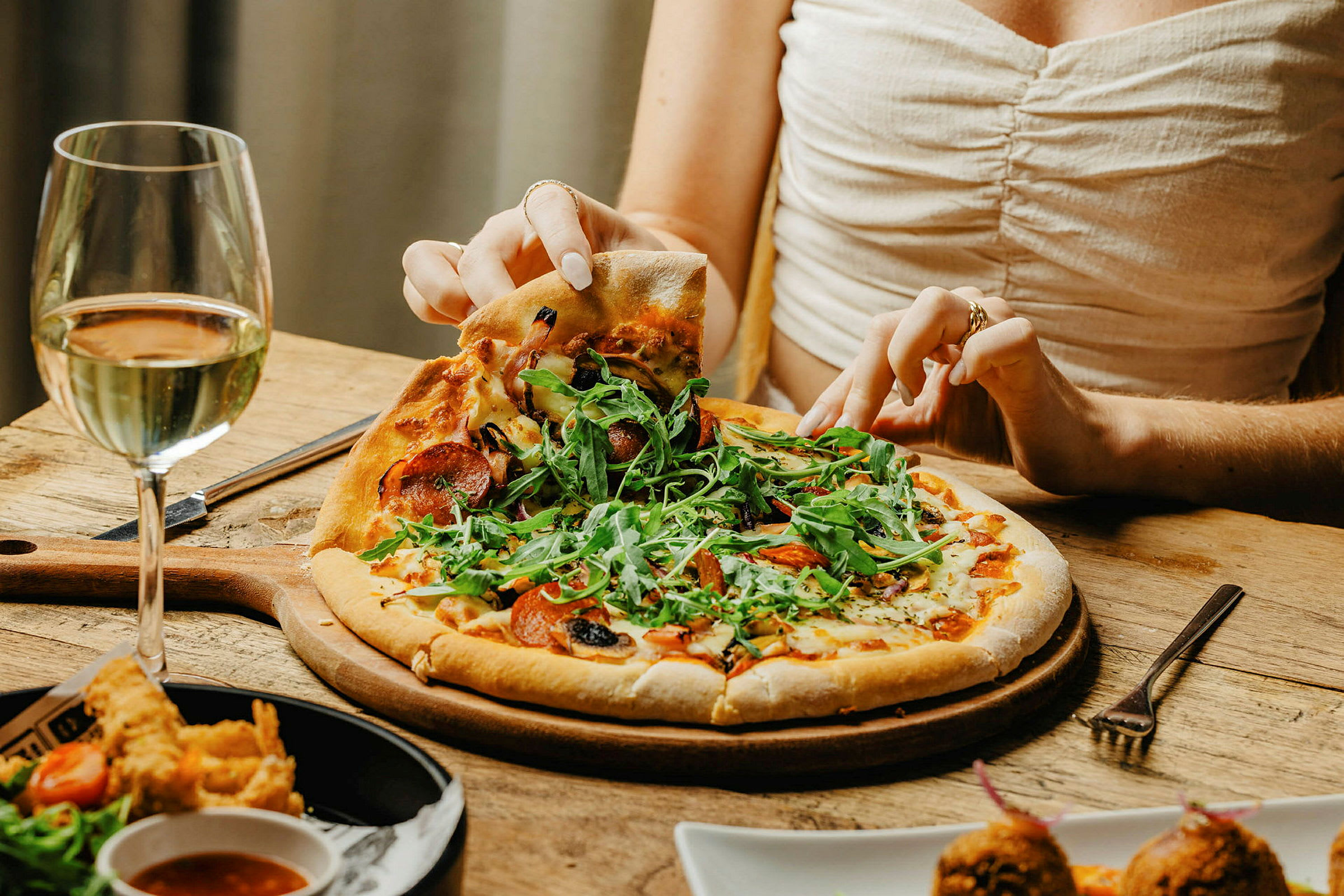 Närbild på en person som tar en bit pizza från en tallrik med en glas vitvin bredvid. (AI-genererad text)