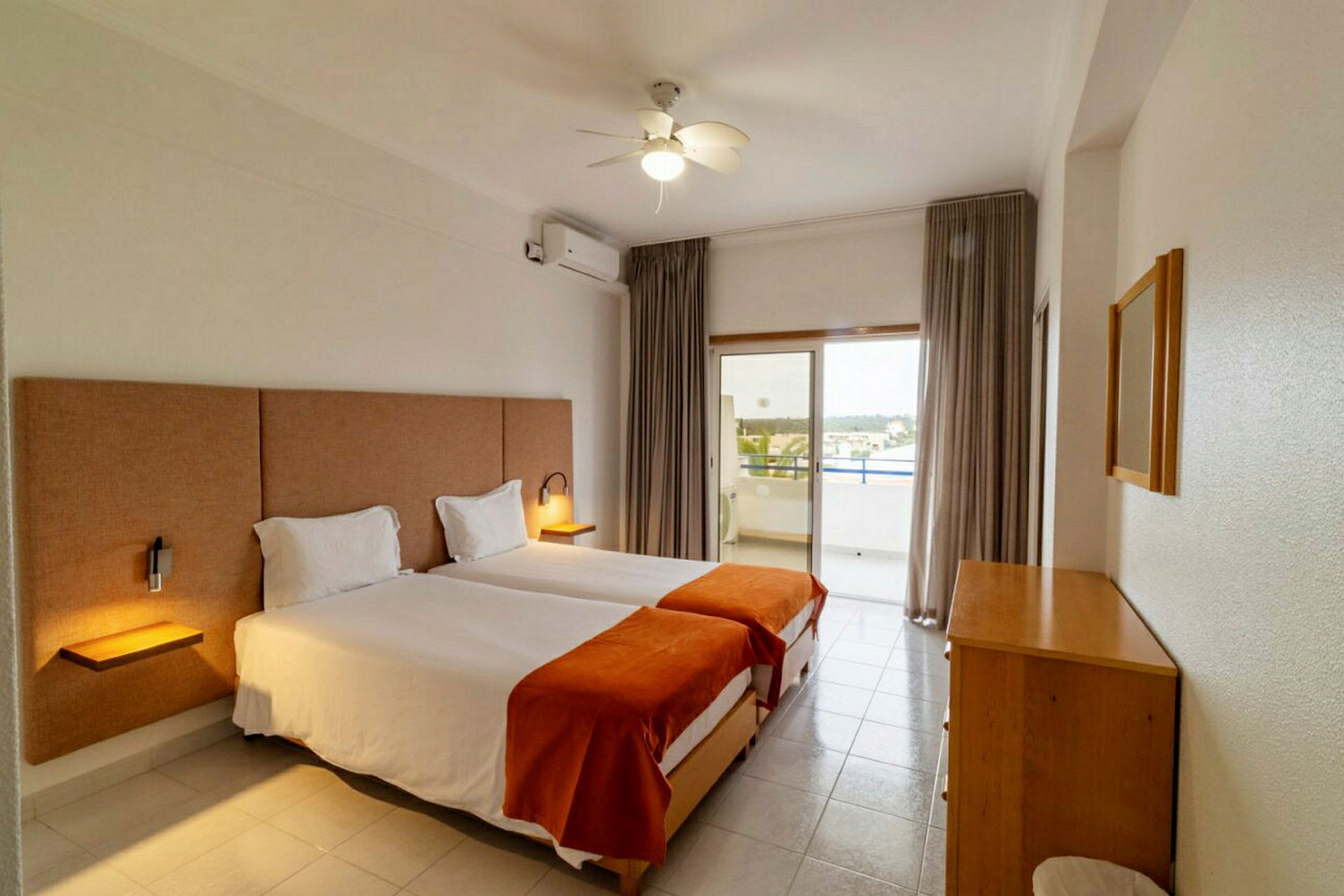 Ett hotellrum med två enkelsängar, orangea plädar och ett stort fönster med balkong. (AI-genererad text)