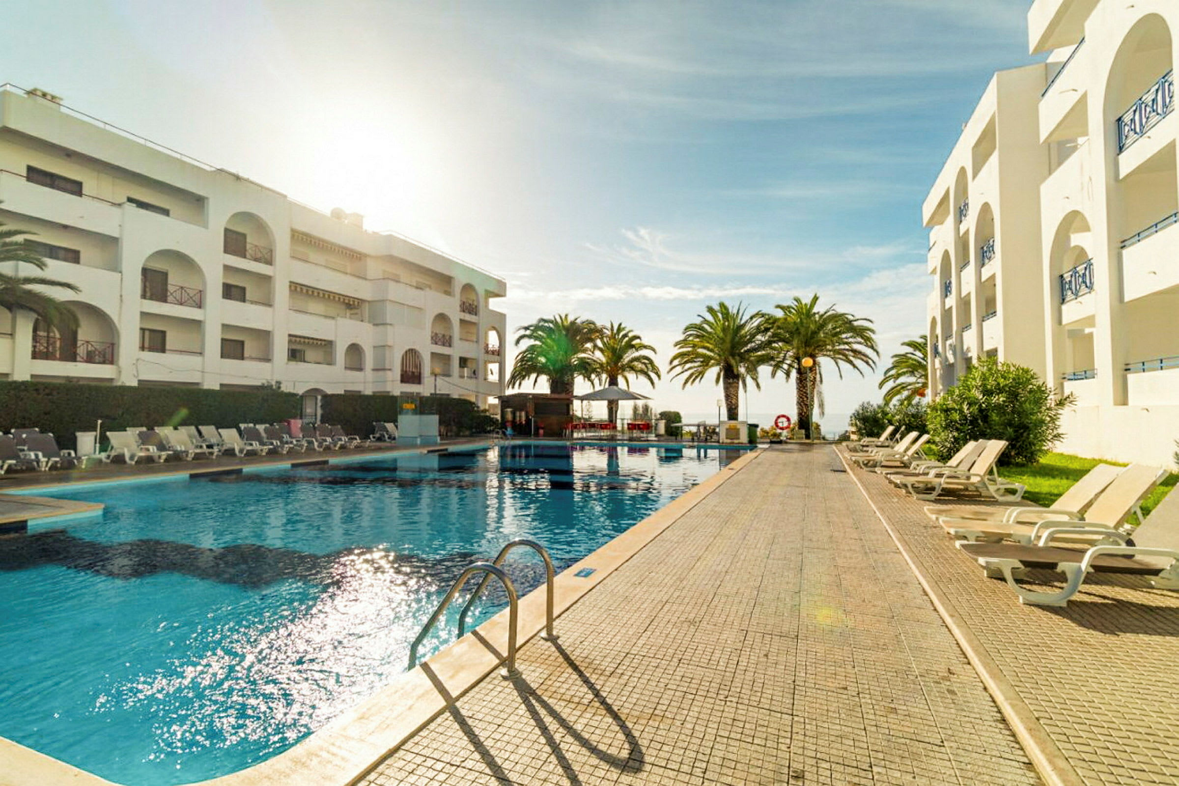 Hotellpool omgiven av solstolar och palmer med en byggnad i bakgrunden. (AI-genererad text)