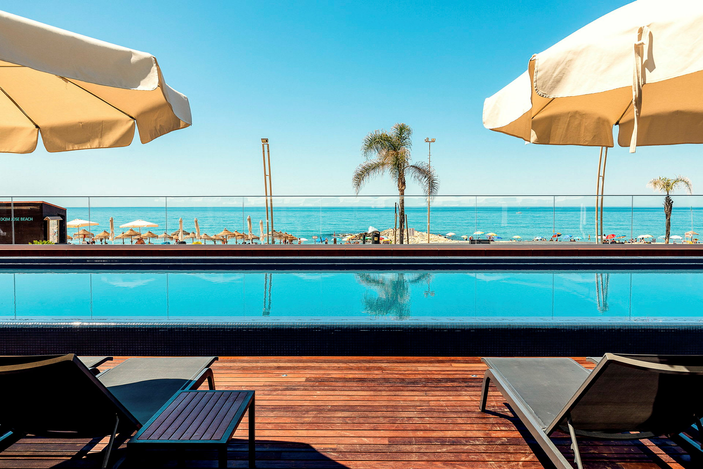 Bild på en pool med havet och strandparasoller i bakgrunden. (AI-genererad text)