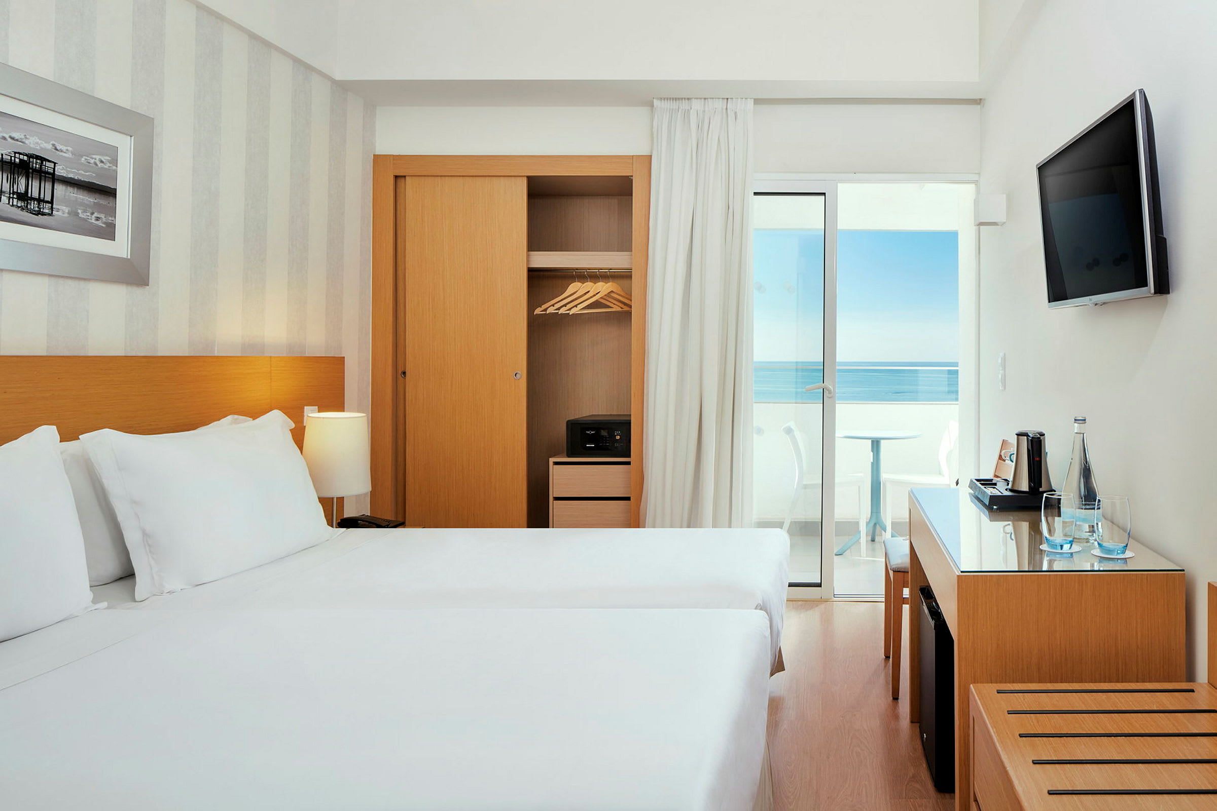 Ett hotellrum med två sängar, garderob, skrivbord och utsikt mot havet från en balkong. (AI-genererad text)