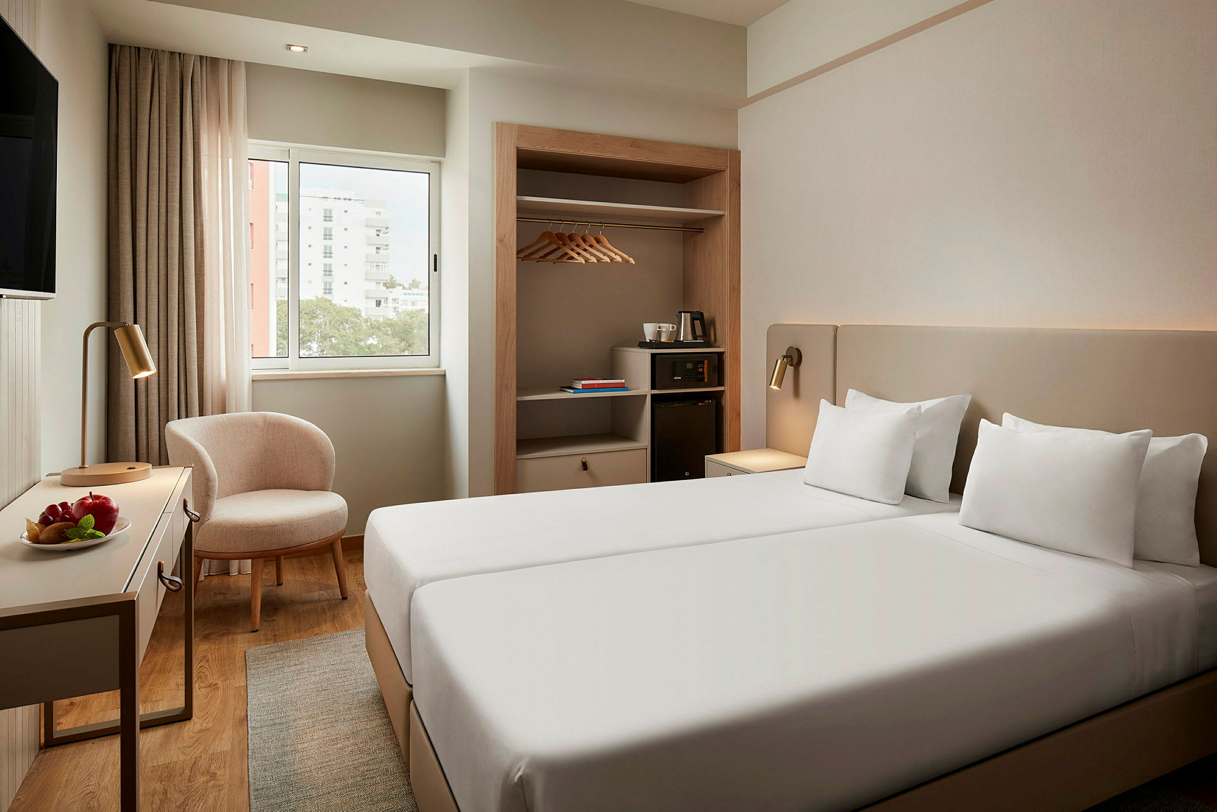 Ett hotellrum med två enkelsängar, ett fönster, en stol, ett bord med fruktskål, och en garderob med hängare. (AI-genererad text)
