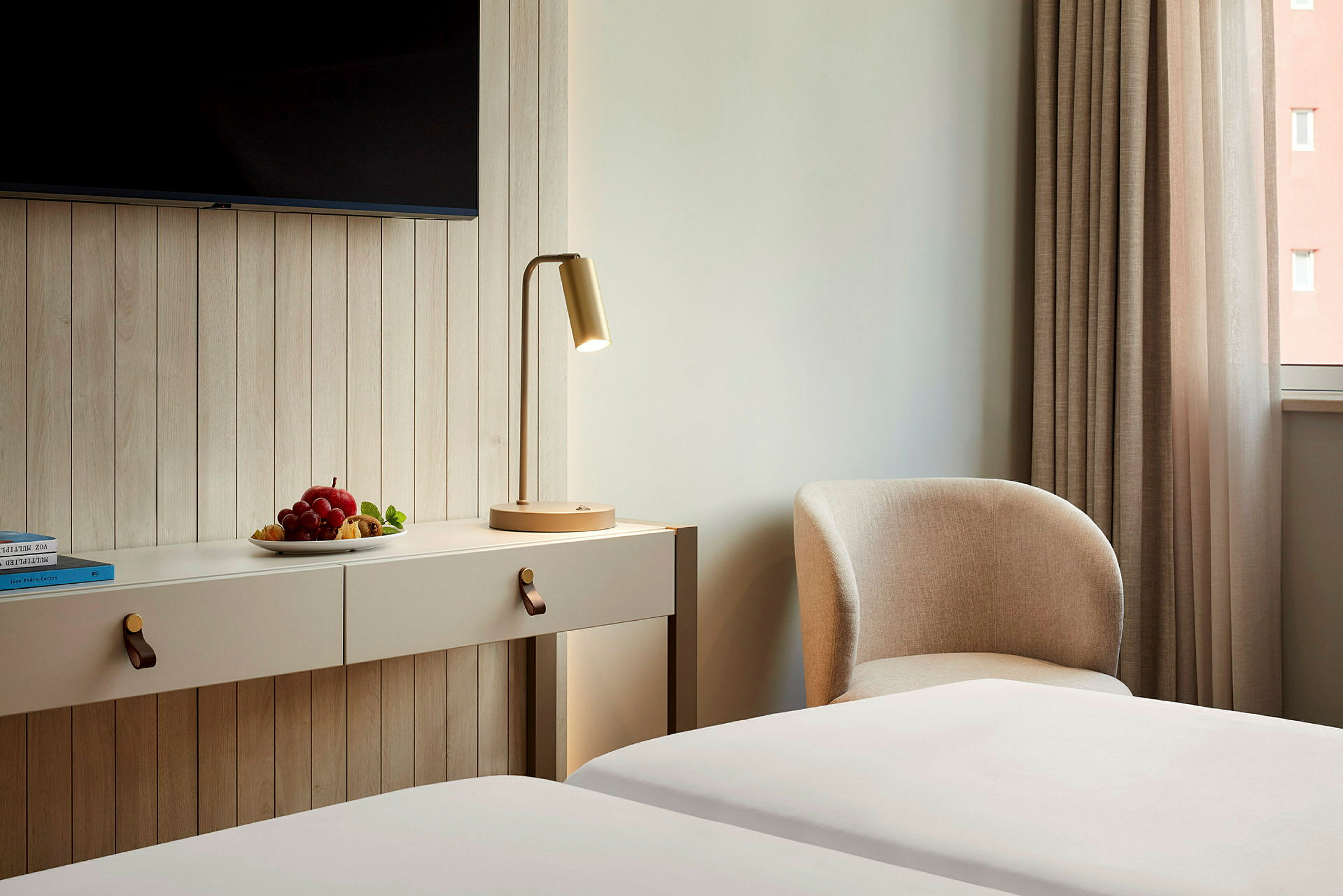 Ett hotellrum med en väggfast möbel, en bordslampa, och en fåtölj bredvid en gardin samt en tallrik med frukt. (AI-genererad text)