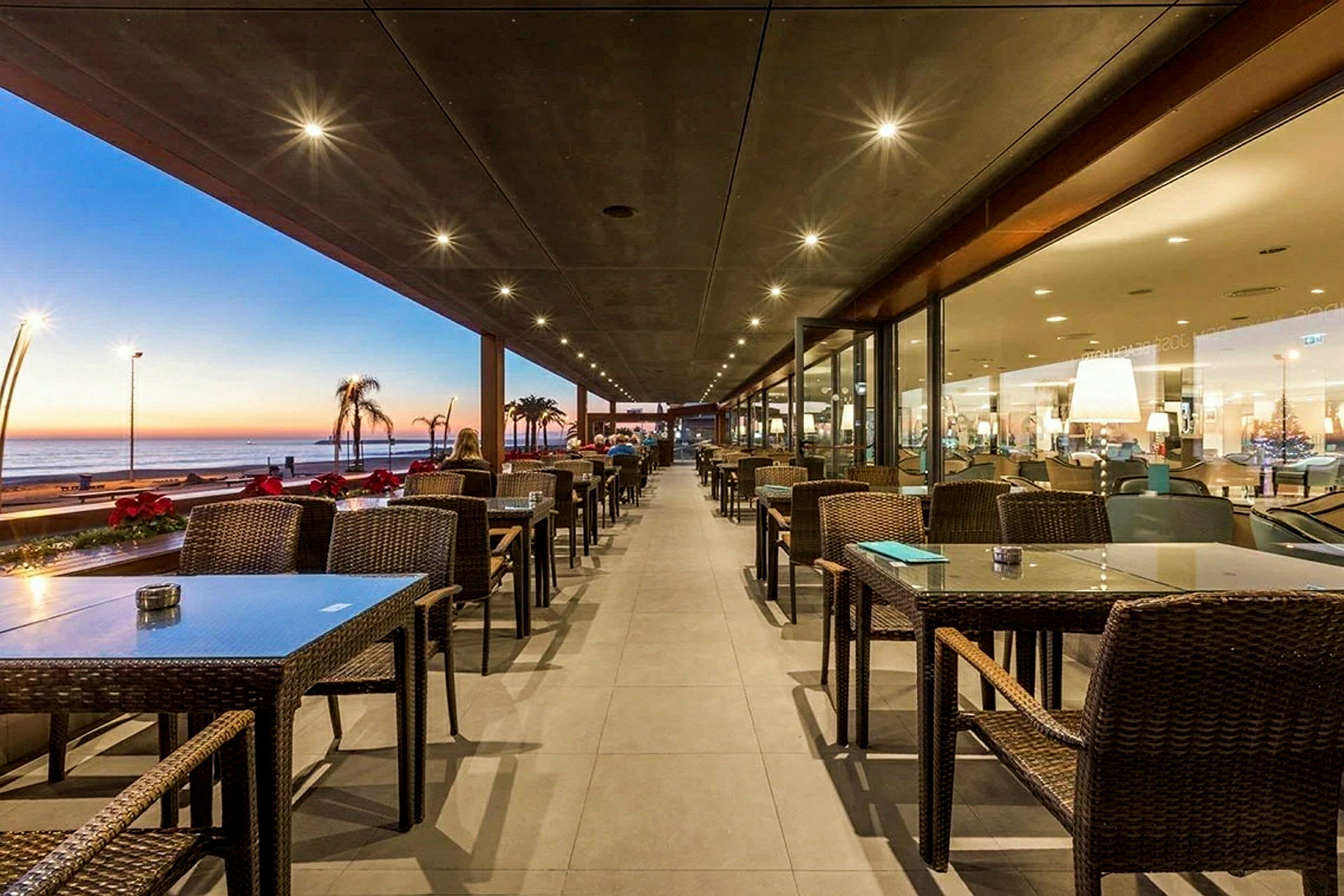 Utomhusrestaurang med uppställda bord och stolar, samt utsikt över havet under solnedgång. (AI-genererad text)