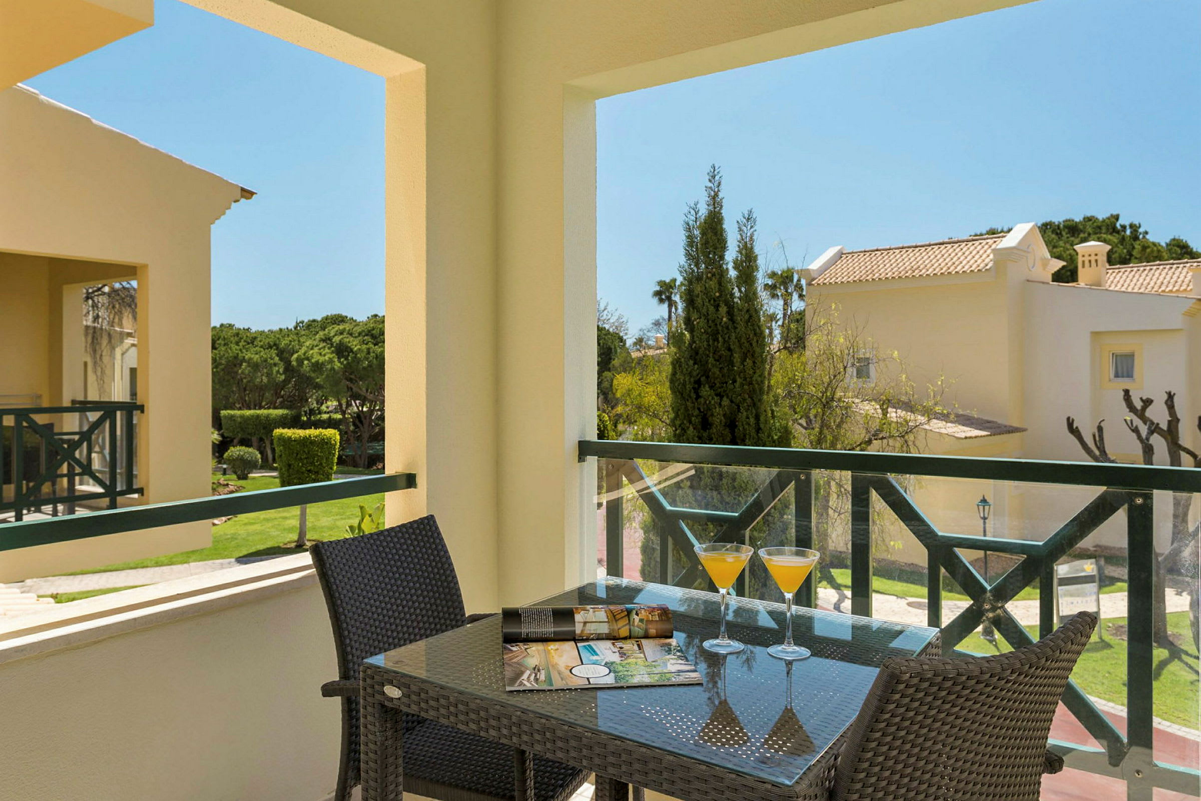 Utsikt från en terrass med glasbord, drinkar och broschyr, omgiven av grönt område och byggnader. (AI-genererad text)