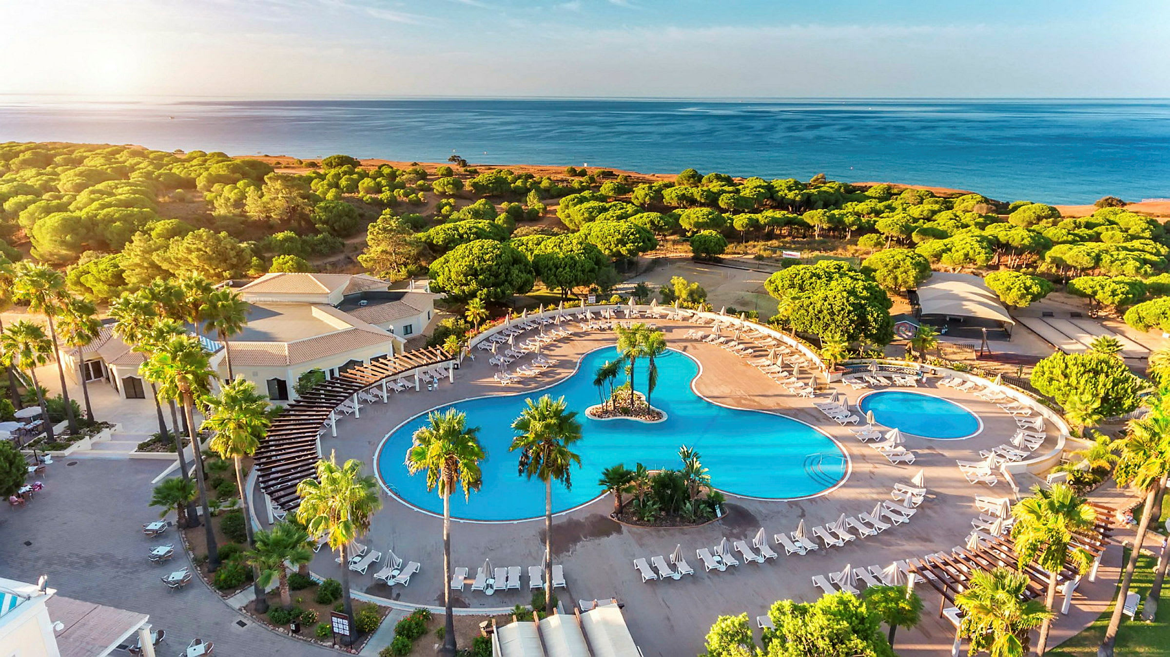 Flygfoto av ett hotellområde med stora pooler, solstolar och omgivande grönska nära stranden. (AI-genererad text)