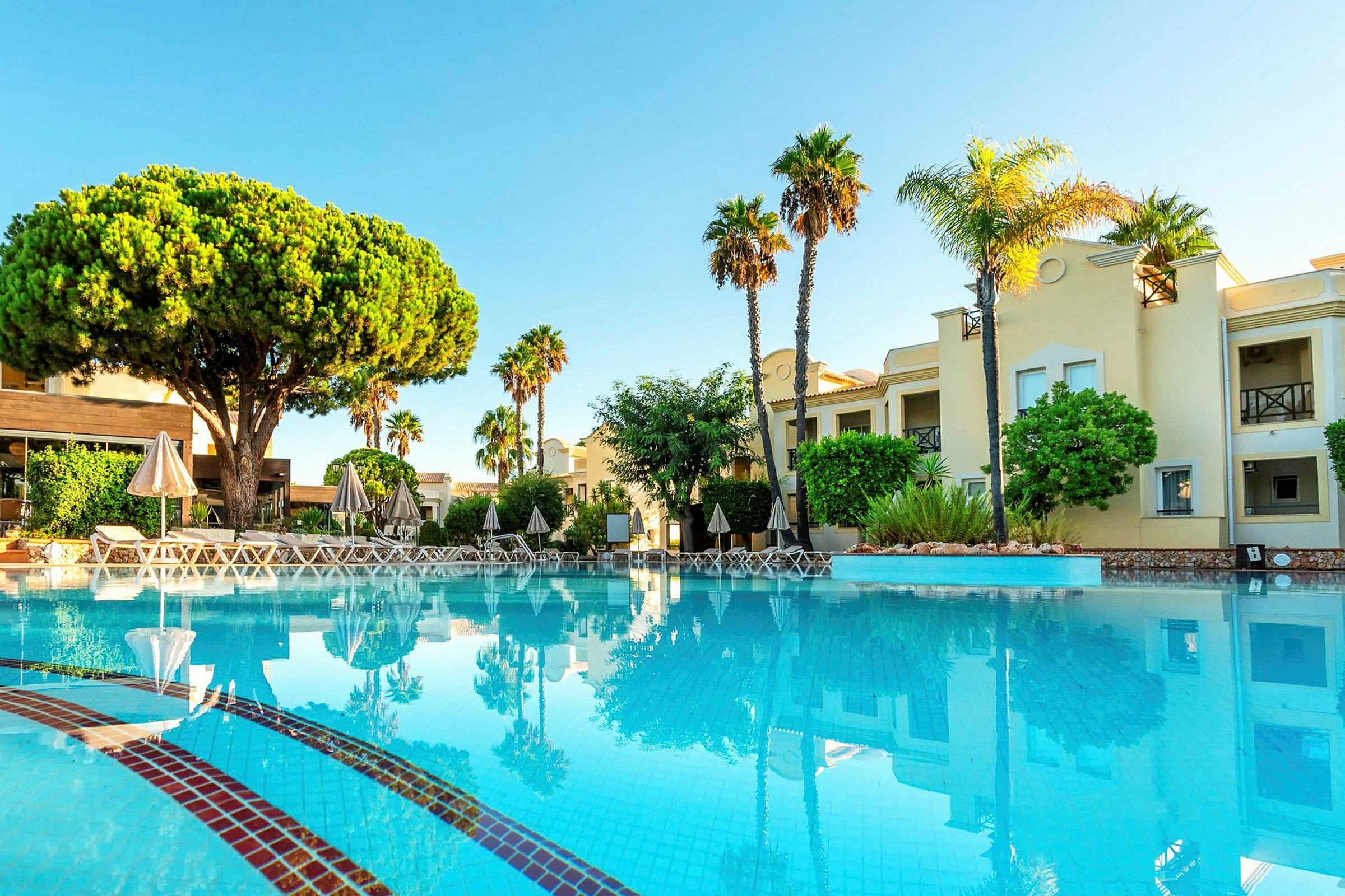 En pool framför en byggnad med träd och solstolar. (AI-genererad text)
