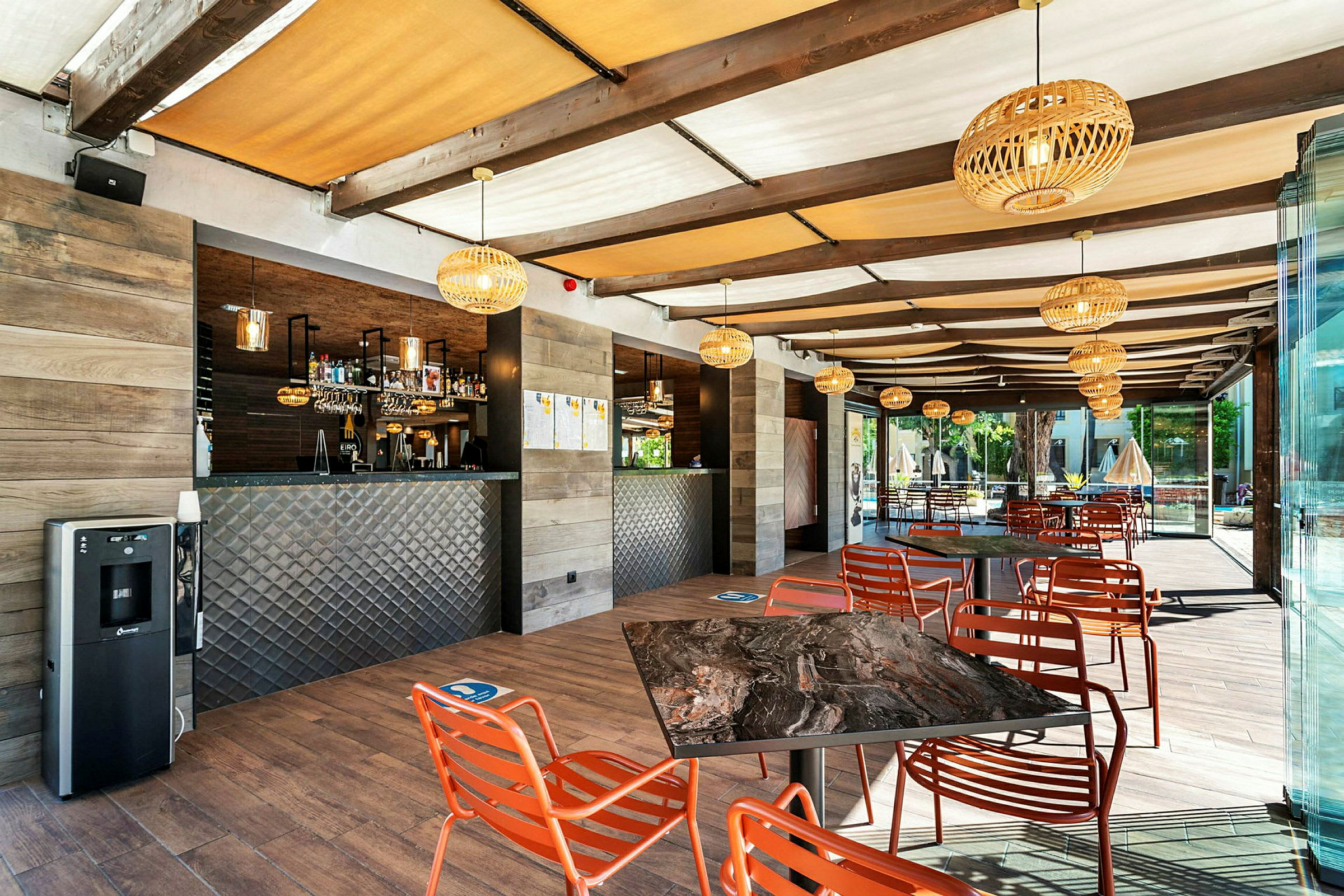 En utomhusrestaurang med bord och stolar i orange. En bar är synlig i bakgrunden. (AI-genererad text)