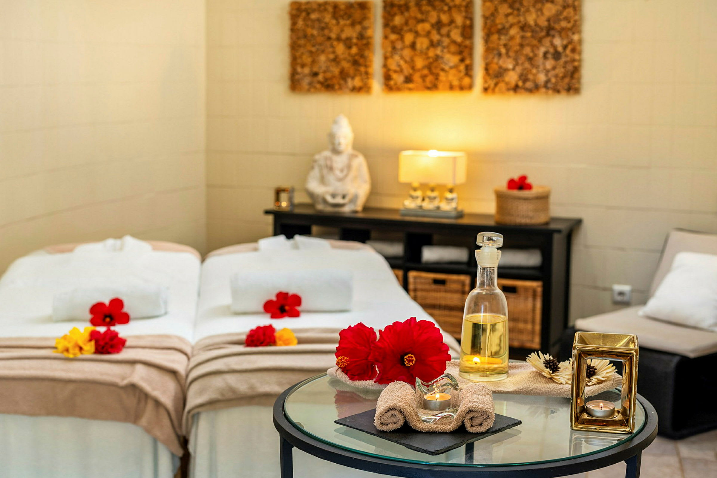 Ett spa-rum med två bäddar, dekorerat med handdukar och blommor. En glasbord med aromatiska oljor och ljus framför bäddarna. (AI-genererad text)
