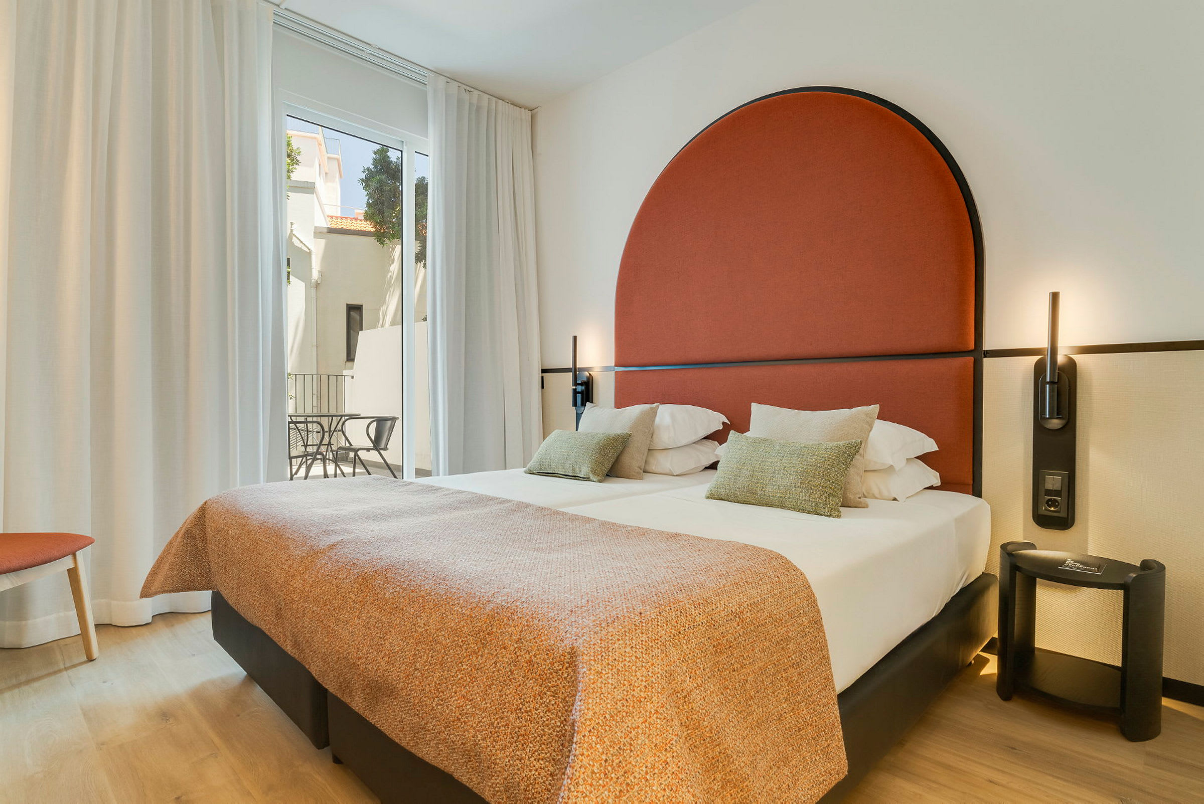 Modern hotellrum med dubbelsäng, röd huvudgavel och stora fönster mot en balkong. (AI-genererad text)